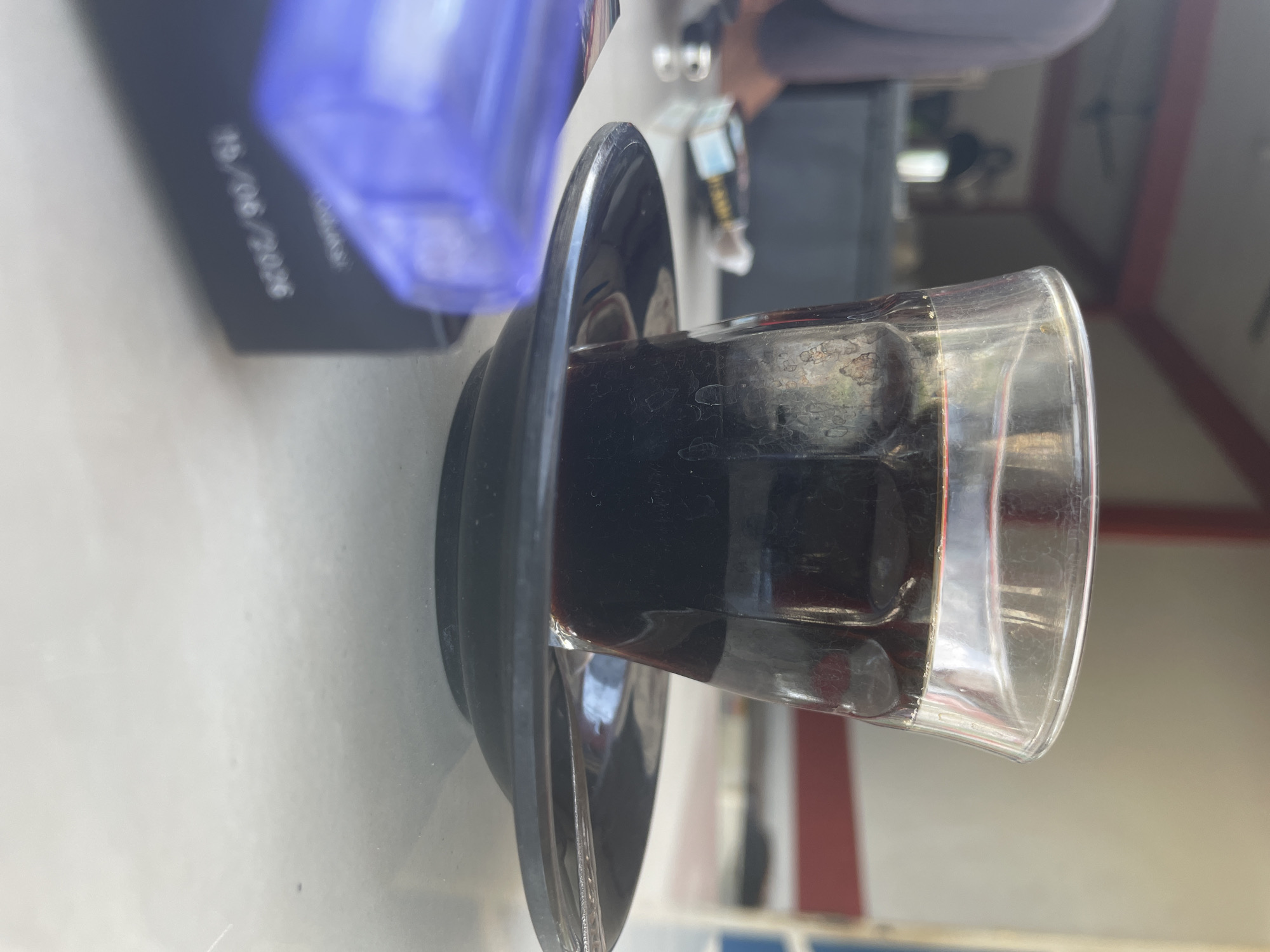 A low-angle, close-up shot shows a clear glass of dark liquid, likely coffee or tea, sitting on a small black saucer. The glass has some water spots and is slightly grimy. In the foreground, a blurred, translucent purple object, possibly a lighter, is visible. To the left of the glass, a cigarette pack is also visible. The background is out of focus, showing hints of an outdoor setting with a roof structure supported by red beams, and the silhouette of a person. The scene appears to be outdoors, possibly at a cafe or roadside stall in Muliengmanyang, Indonesia, given the cultural context often associated with such beverages and setting. The image is taken during daylight.
