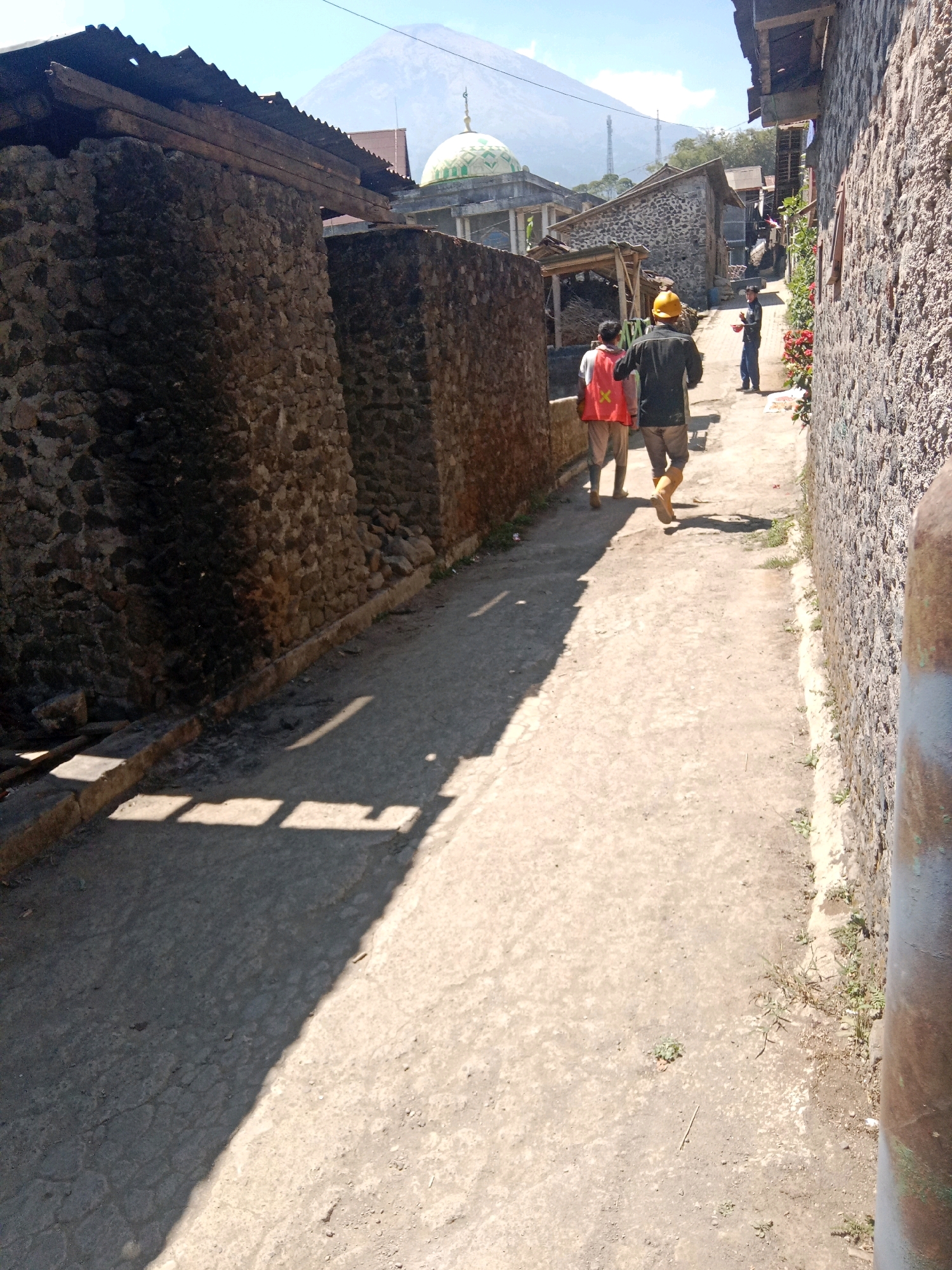 The image shows a narrow, paved pathway in Jurangjeru, Indonesia, with rough stone walls on either side. In the background, a mosque with a green dome is visible, and a mountain looms beyond it. Three men are walking away from the camera down the path. The man in the foreground wears a yellow hard hat, a dark jacket, and light-colored pants. To his left, another man wears a bright orange vest over a dark shirt and light pants. A third man, further down the path, is wearing a blue shirt and dark pants. The sun casts long shadows across the pathway, indicating it is likely midday. The environment appears to be rural or a small village, with traditional stone construction.