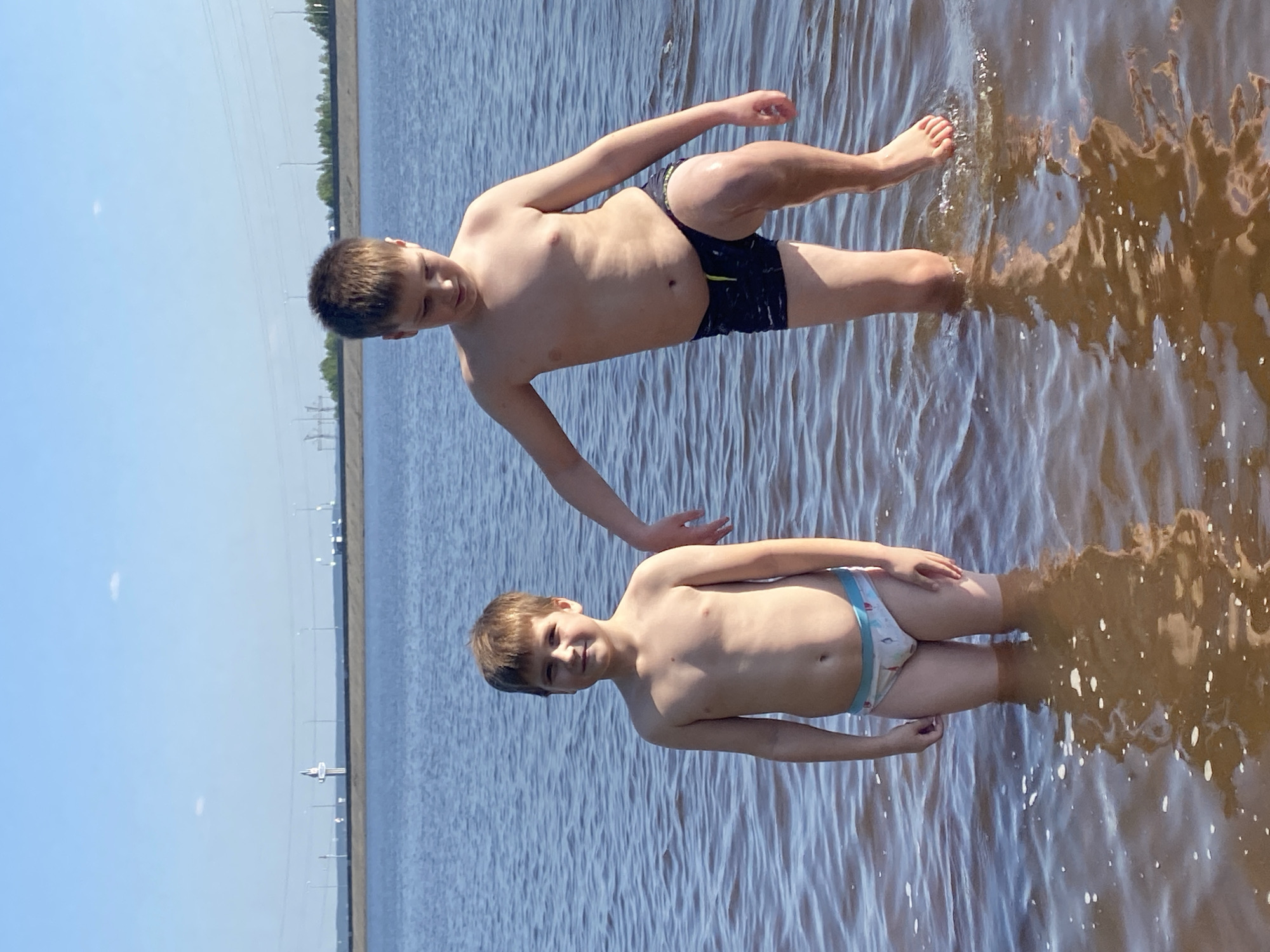 Two shirtless young boys are standing in shallow water. The boy closer to the viewer is wearing light-colored swimwear with a blue stripe at the waist and a design. The other boy is wearing dark swimwear. The water is slightly rippled, with reflections. The background shows a flat horizon with a levee and utility poles, suggesting a location near Chaikovsky, Russia. The sky is blue and sunny.