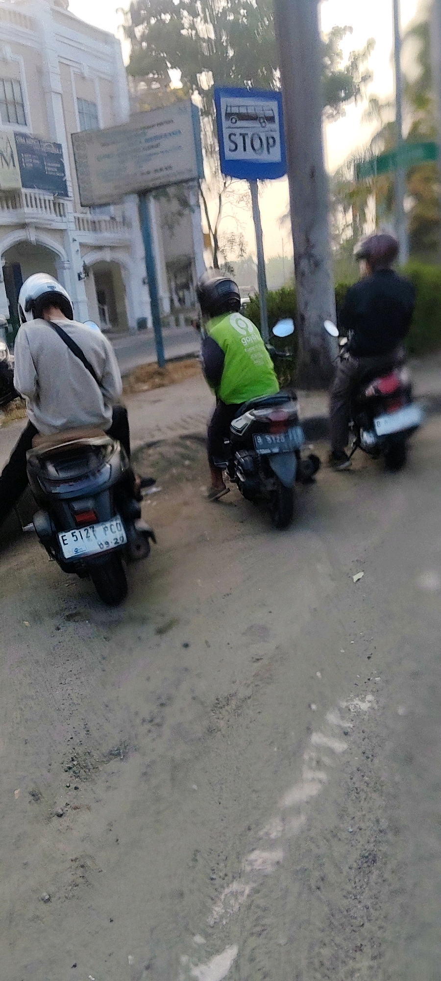 In Pasirsari, Indonesia, three individuals are observed on motorcycles at a bus stop. A person in a white helmet, wearing a grey long-sleeved shirt and dark pants, sits on a motorcycle in the foreground, facing away from the camera. To their right, another person, also facing away, is dressed in a bright green Gojek jacket and dark pants, seated on a motorcycle. A third individual, wearing a flat cap and dark clothing, is on a motorcycle further to the right and slightly behind the other two, also facing away. A blue bus stop sign with the word "STOP" is visible behind the motorcycles. The setting appears to be a street with a textured surface, possibly asphalt or dirt, and a building with arched windows is present in the background. The lighting suggests it is either early morning or late afternoon.