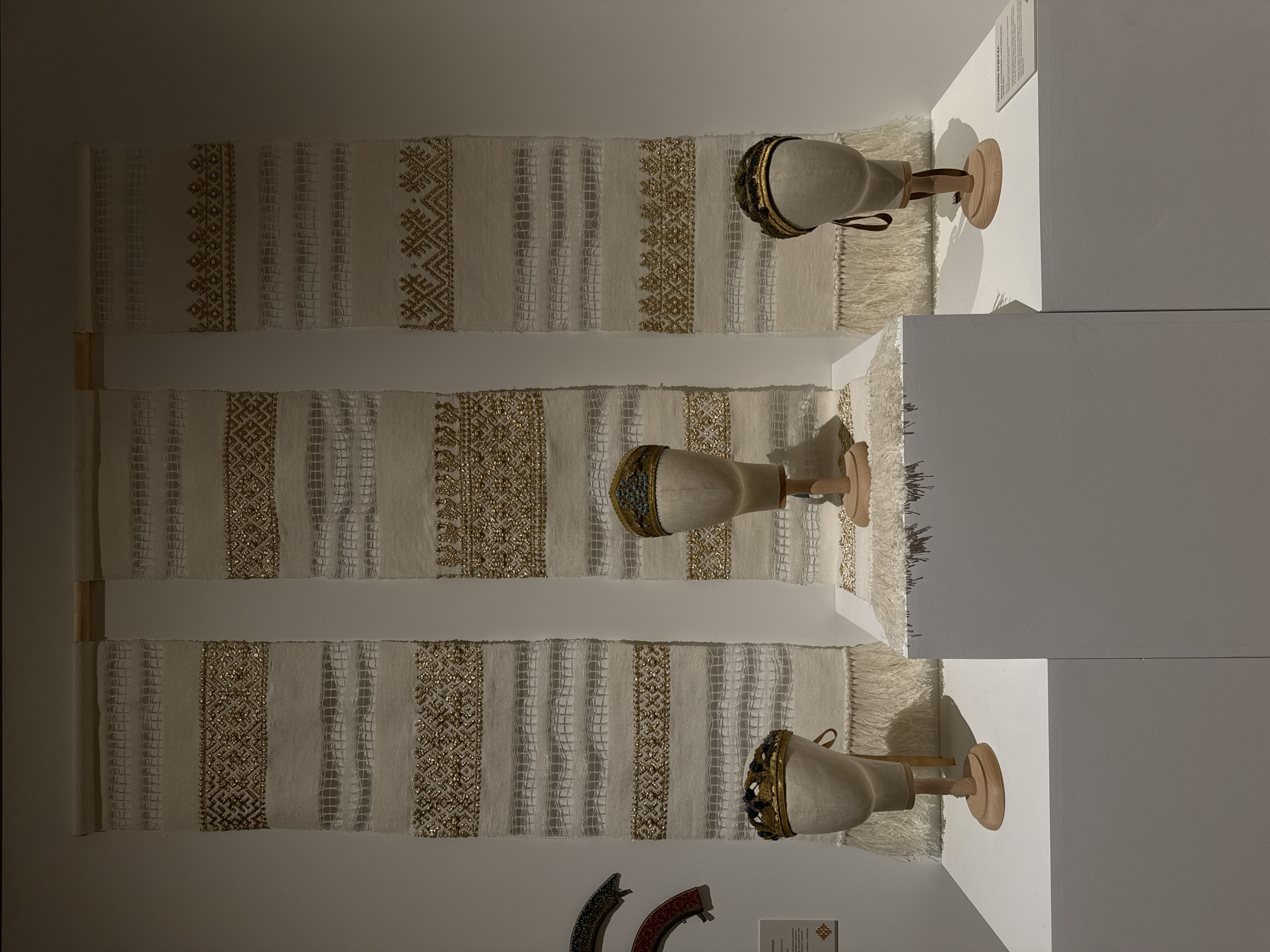The image captures an interior exhibition space, likely a museum or gallery, showcasing traditional embroidered textiles and headwear. Three large, white, woven textiles with intricate gold-colored embroidery hang vertically against a light-colored wall. Each textile has a fringe at the bottom. Mounted on pedestals next to each textile are three headpieces displayed on simple, light-colored mannequins. The headpieces are ornate, with gold accents and embellishments. In the lower part of the image are fragments of other textiles and a small information panel. The environment is clean and well-lit. The location, Moscow, Russia, is not evident from the image alone.