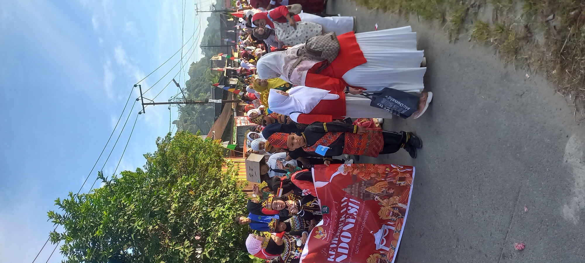 This image depicts a street parade in Samarinda, Indonesia, possibly celebrating Indonesian Independence Day given the banner's inscription "KEMERDEKAAN INDONESIA." A group of children and adults are participating, with some children dressed in traditional attire, including a boy in a black and red outfit with a patterned sash and headwear. A prominent banner held by some participants displays the text "KEMERDEKAAN INDONESIA" and "SDIT NURUL HADI SAMARINDA," indicating the school's participation.
The scene is outdoors on a paved street lined with trees and buildings. Utility poles with power lines are visible in the background, along with distant hills. A few people are carrying bags, including one with a McDonald's logo. The sky is clear blue with some wispy clouds. The overall atmosphere appears festive and communal.