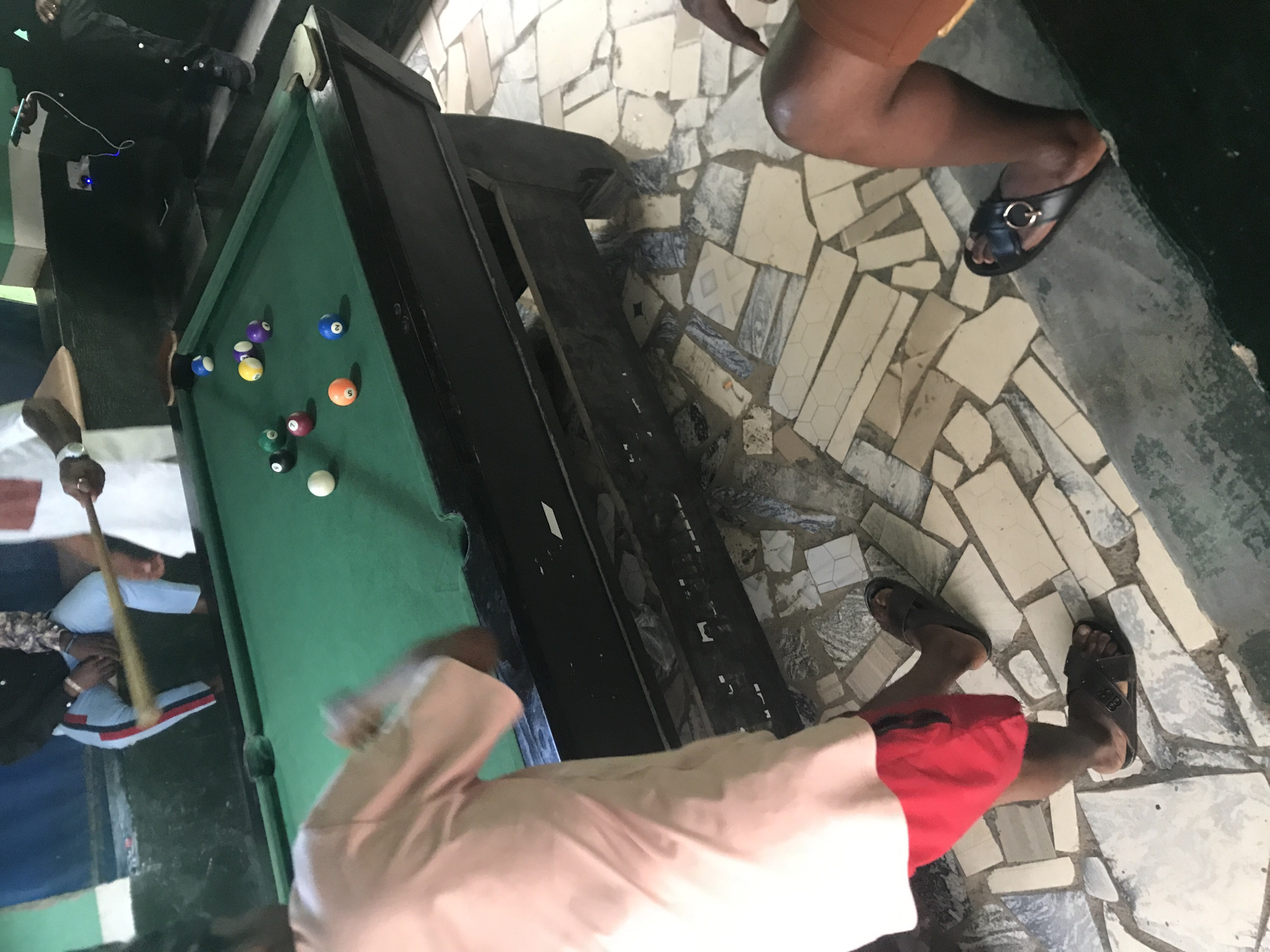 In an indoor setting, a billiard table with green felt and black trim is the central focus. Several billiard balls are scattered on the table, including a white cue ball and various colored balls numbered from 2 to 9. A billiard cue is held horizontally across the table by two unseen individuals, with one hand visible gripping the stick.
In the foreground, a person wearing a light pink shirt and red shorts stands with their back to the camera, their legs partially visible. They are wearing black slide sandals. To the right, another person's legs are visible, wearing brown shorts and black slide sandals with a gold accent. The floor surrounding the table is covered with a mosaic of light-colored and gray rectangular tiles, some with geometric patterns. The background is dimly lit, with hints of a blue wall and a counter. There is also evidence of electrical outlets and wires on the wall.