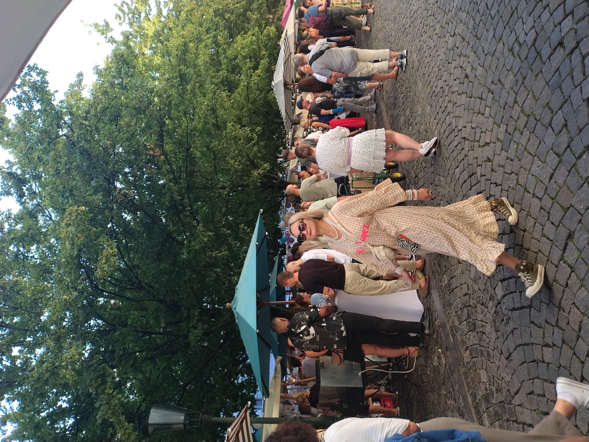The image depicts an outdoor scene at an event, possibly a street festival or market, on a cobblestone pavement. The atmosphere appears to be festive with several people gathered. In the foreground, a woman is walking towards the right, wearing a checkered dress, a pearl necklace, and sunglasses. She is wearing green sneakers. Behind her, other individuals are seen mingling and standing around. One man is wearing a black shirt with white graphics, another is wearing a black t-shirt, and a woman is wearing a white dress with a floral pattern. In the background, there are trees, umbrellas providing shade, and market stalls or stands. The lighting suggests it is daytime, possibly late afternoon or early evening. The presence of trees and the architectural style of the lamppost suggest a European city, and while specific details to confirm Prague, Czechia are not overtly present, the scene is consistent with urban outdoor gatherings found in such locations.