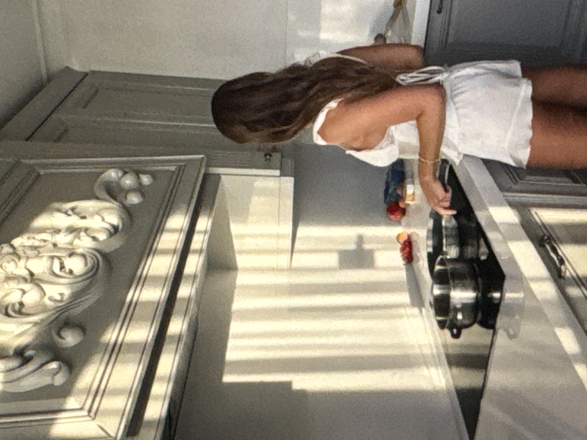 A person is standing in a kitchen with light streaming in from a window, casting shadows across the scene. The person, with long dark hair, is wearing a white outfit. In front of them is a stovetop with pots and pans, suggesting they may be cooking. The kitchen appears to have white cabinets and a light-colored countertop. To the left, a large ornate cabinet door is visible, decorated with intricate carvings. The overall impression is a bright and domestic scene, possibly in the morning or late afternoon given the angle of the light. No visible text is present in the image.
