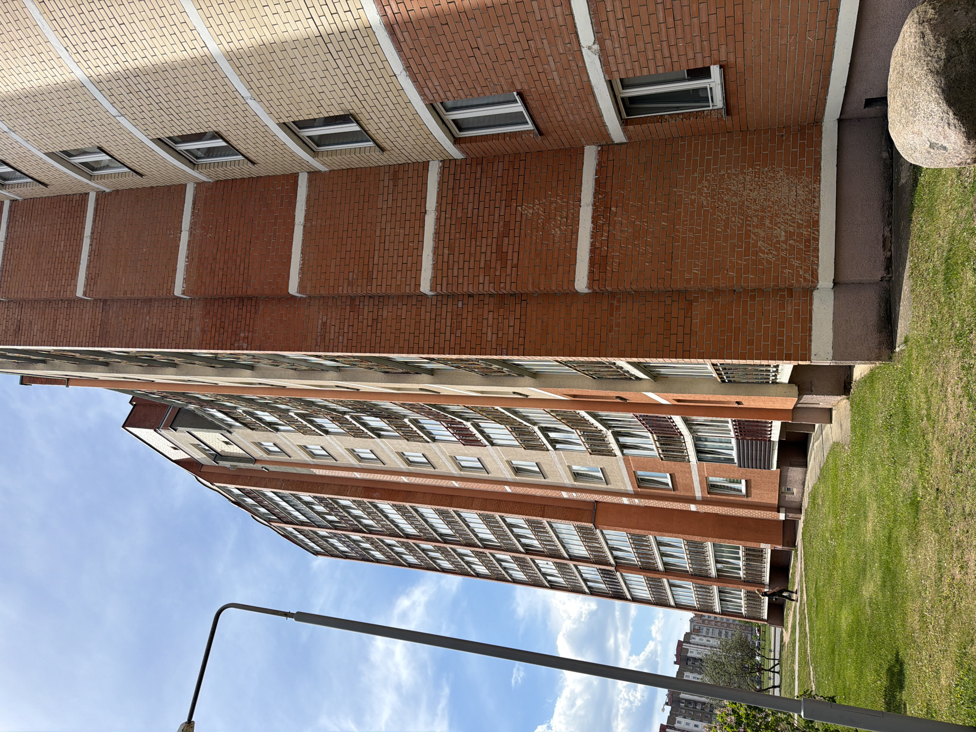 The image shows a tall brick apartment building under a bright blue sky with scattered clouds. The building has numerous windows and a recurring vertical architectural element that alternates the colors of the brickwork, including white detailing. The building extends out of frame on the left side. Below the building is a grassy area with a light post that has a small brass bulb. In the background, more buildings are visible. The image appears to be rotated counterclockwise. The location of the image is Unknown, Unknown.