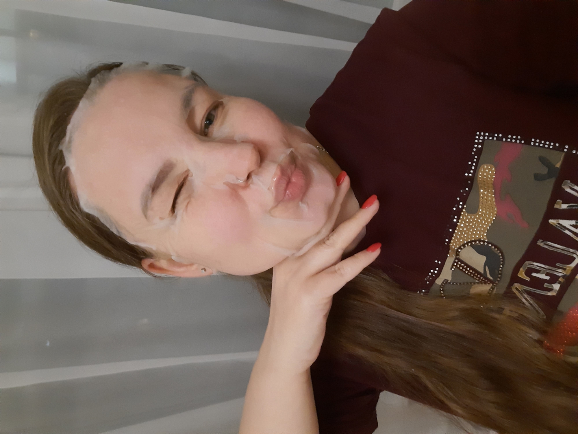 A person with a sheet mask on their face is winking and pursing their lips. They are wearing a maroon shirt with embellishments. The background appears to be a gray curtain. There is no information in the image to suggest that this photo was taken in Izhevsk, Russia.