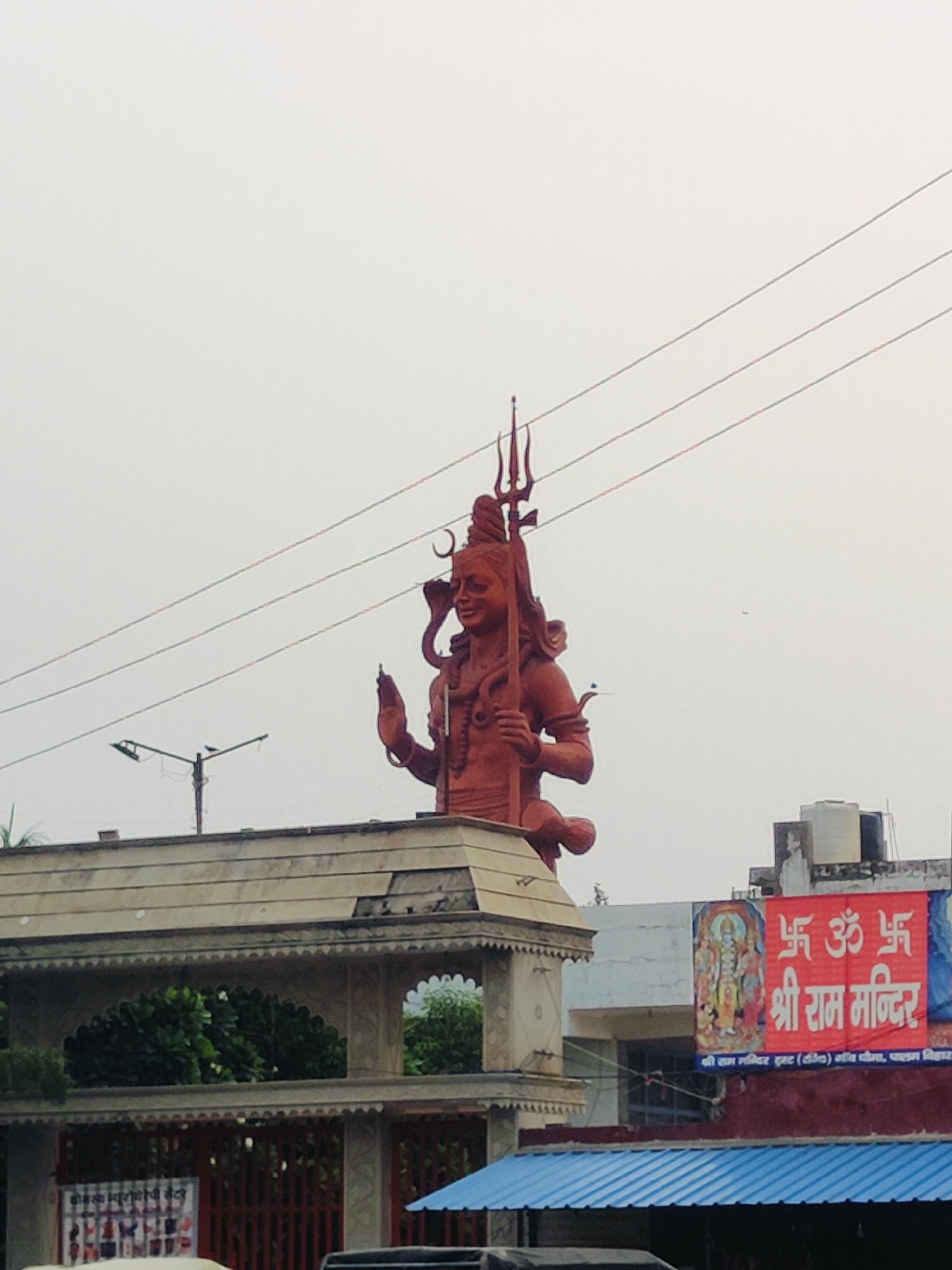 This image captures a large, reddish-brown statue of a Hindu deity, likely Shiva, prominently displayed outdoors against a pale, overcast sky. The statue is depicted with multiple arms and holds a trident. It appears to be situated in Gurgaon, India, as indicated by the signage in the lower right corner which reads "श्री राम मन्दिर" (Shri Ram Mandir). The statue is positioned behind an architectural structure resembling an archway or gate. In the background, there are buildings, water tanks, and electrical wires. A red banner with religious symbols and text is hung on one of the buildings. The overall scene suggests a public space, possibly a temple complex or a significant landmark. There are no visible people actively engaged in any interaction with the statue or other elements in the scene. The environment appears to be an urban setting in Gurgaon.