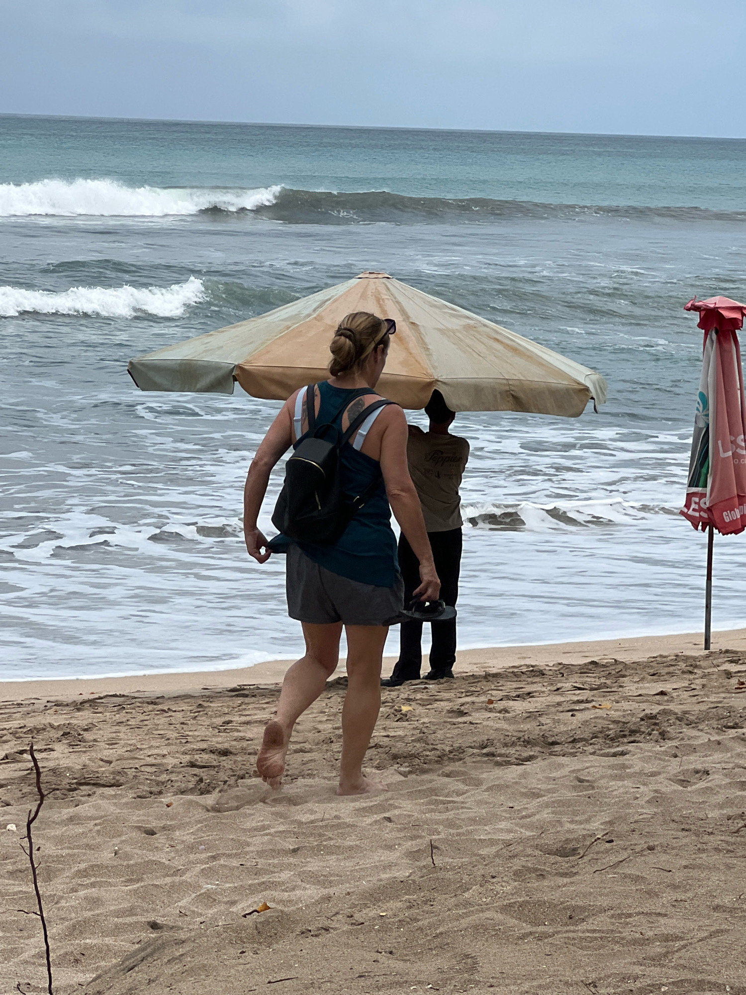 In Kuta, Indonesia, a woman with blonde hair pulled back in a bun is walking barefoot on a sandy beach. She is wearing a dark tank top, grey shorts, and a black backpack. In front of her, a person wearing a tan t-shirt and dark pants stands under a tan umbrella, holding a red umbrella to their side. The ocean with white-capped waves is visible behind them, and the sky is overcast. Another red umbrella is propped up on the right side of the frame. The overall scene depicts a cloudy day at the beach.