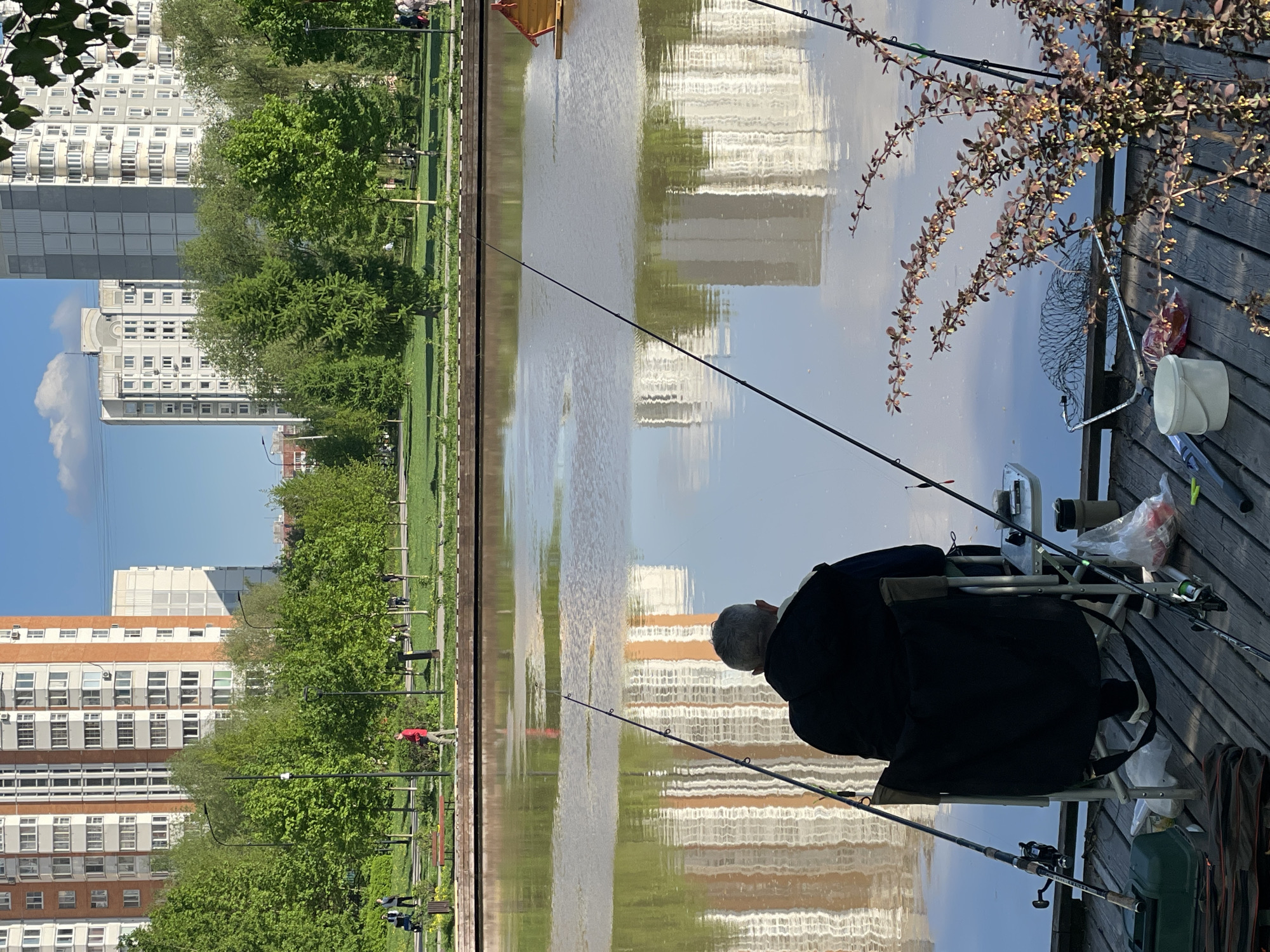 An image shows a man fishing from a wooden platform on a body of water in a park setting. The man is seated in a folding chair, with two fishing rods extended into the water. Fishing equipment is placed around him on the wooden platform. Across the water, a city park with trees and walkways can be seen, with tall apartment buildings visible in the background. The water surface reflects the buildings and the sky. The photograph shows a day scene with clear, sunny weather. The location is Moscow, Russia.
