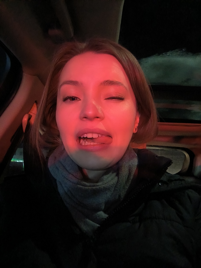 A fair-skinned woman, illuminated by a red light, winks with her right eye and sticks her tongue out. She wears a gray scarf and a dark, puffy jacket. The setting appears to be inside a car at night, evidenced by the dark exterior and the car's interior elements visible around her.