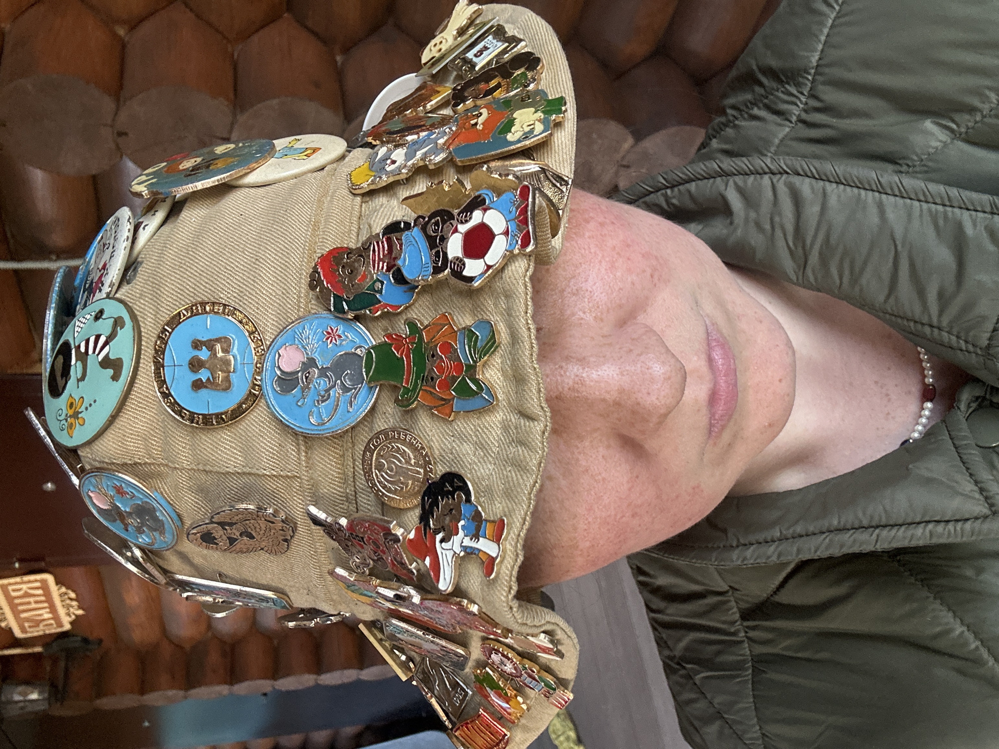 A close-up shot shows a fair-skinned person with freckles wearing a light brown baseball cap covered with numerous pins. The pins feature various cartoon characters and symbols, including a soccer ball, animals, and people. The person is wearing a green quilted jacket and a string of pearls. The background includes a wooden wall and a sign with Cyrillic text, potentially indicating the location. While the image is not specific enough to pinpoint the exact spot, the presence of Cyrillic text suggests that the photo was taken in Russia, likely in a location frequented by tourists or with a unique cultural offering. Given the prompt, it is possible that the location may be in the city of Nizhny Novgorod.