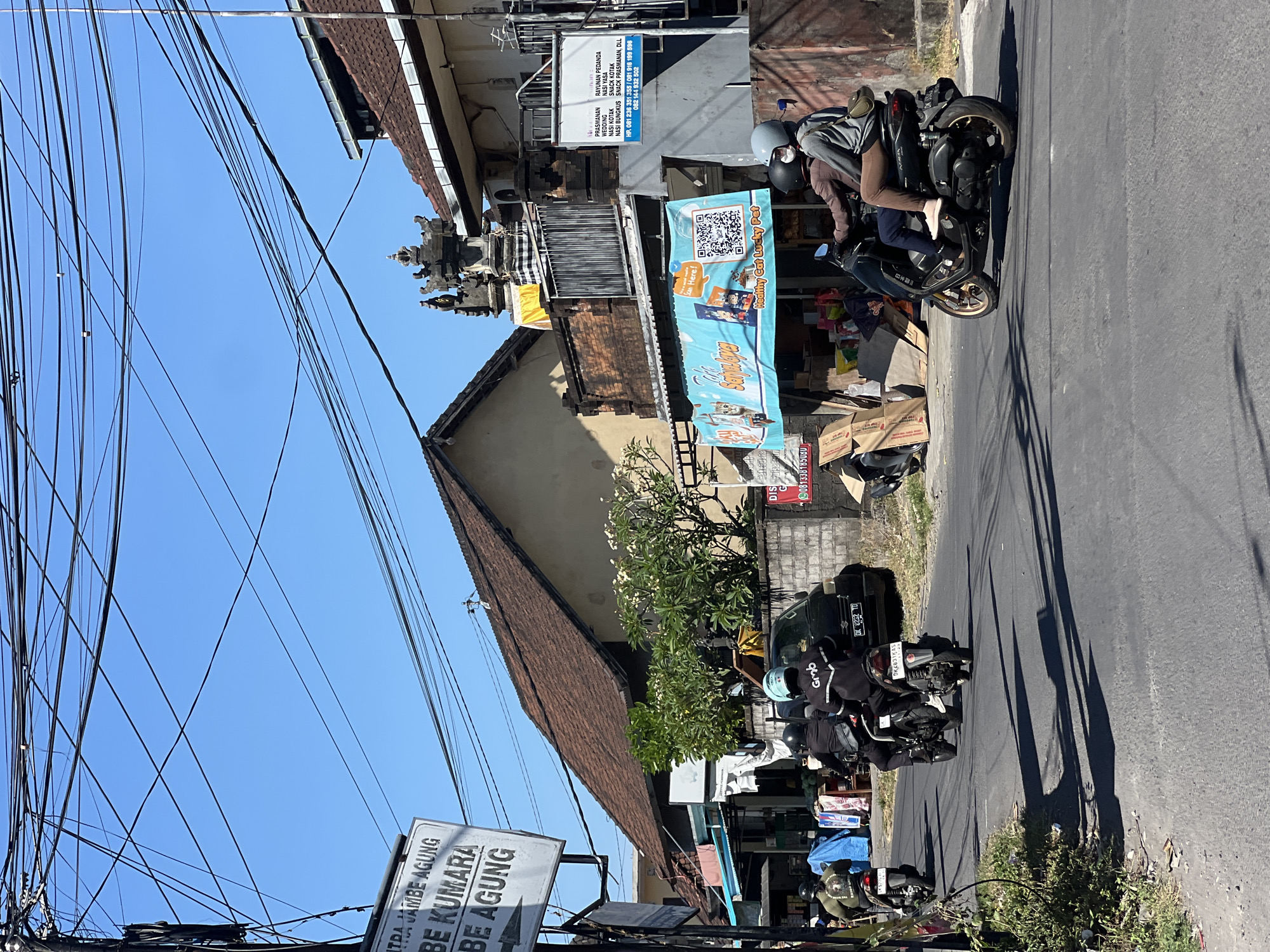 The image depicts a street scene in Sukawati, Indonesia, characterized by a clear blue sky and numerous overhead electrical wires. The foreground shows an asphalt road with shadows cast by the overhead wires. Several motorcycles are visible on the road. Two individuals are riding on a black scooter in the right lane, with the rider wearing a helmet and a brown jacket, and the passenger wearing a helmet and a grey sweatshirt. To the left, another motorcycle with two riders, both wearing helmets, is traveling in the same direction. Further ahead, a black car is parked on the side of the road.
Buildings line the street, featuring traditional Balinese architecture with red tile roofs and ornate stone carvings. One building displays a large blue banner with text and an image of cats, advertising "Healthy Cat Lucky Pet." Another building has a white sign with Indonesian text listing services and phone numbers. A signpost on the left side of the image shows directional information for "JEMBE AGUNG," "BE KUMARA," and "BE AGUNG." Vegetation, including trees with white flowers, is present between some of the buildings. Stacks of cardboard boxes are visible near one of the shops. The overall impression is a typical urban street environment in Bali, with a mix of residential and commercial properties and active street life.
