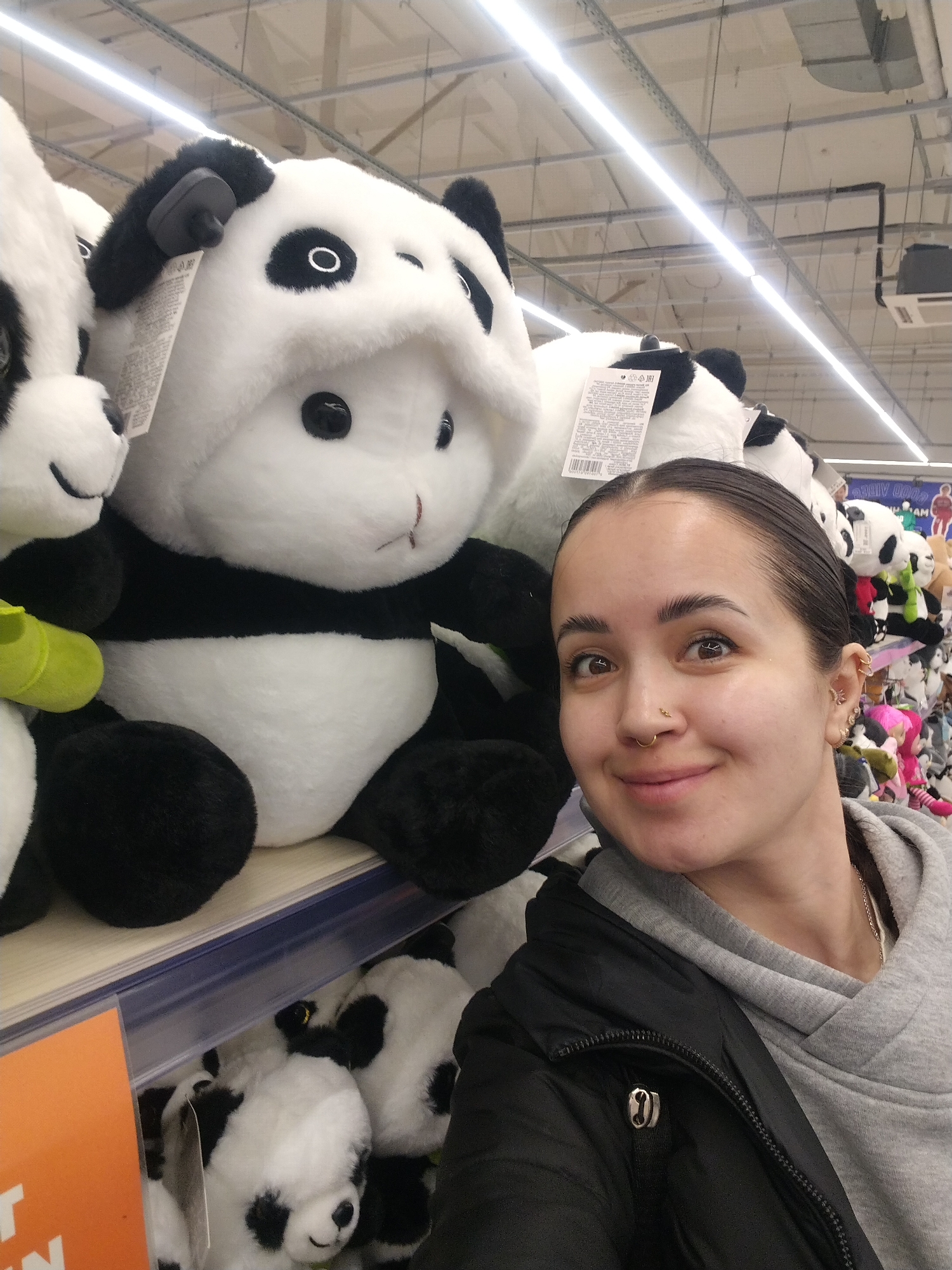A woman is taking a selfie in front of a shelf filled with panda plush toys. She is wearing a grey hoodie and a black jacket. She has her hair pulled back and has multiple piercings in her left ear and a nose ring. The pandas are arranged on multiple shelves, with more pandas visible in the background. The location appears to be a retail store, as indicated by the overhead lighting and shelving units. The city and specific location are unknown.
