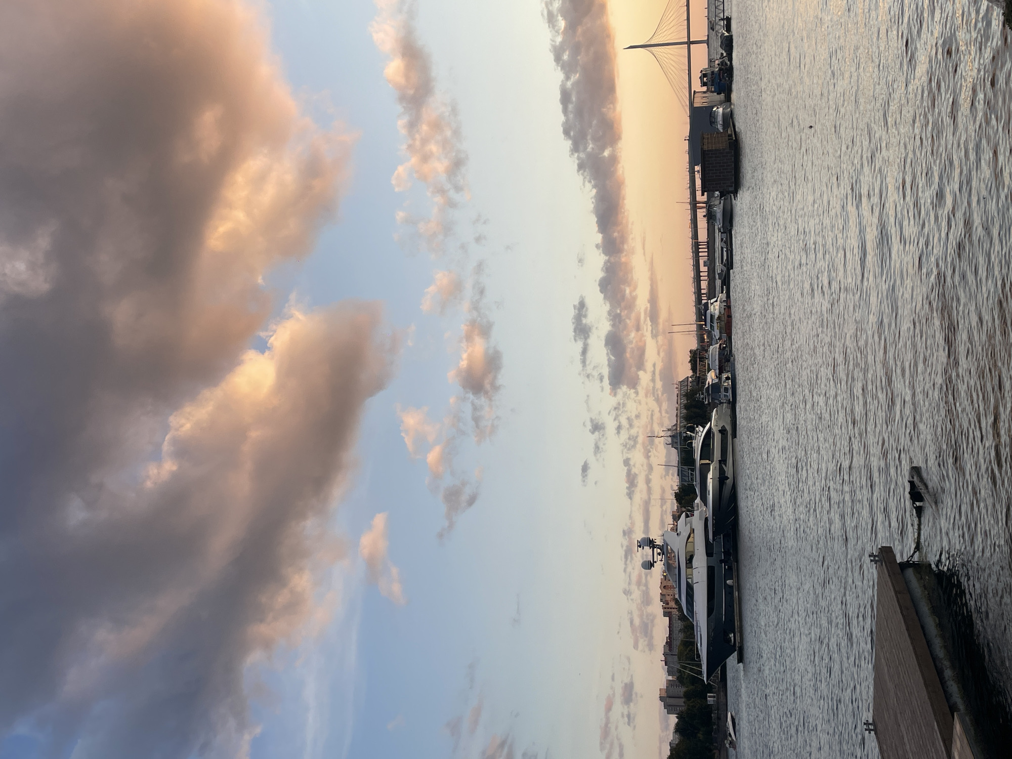 The image captures a serene waterfront scene in Saint Petersburg, Russia, during a sunset. The sky is painted with soft hues of orange, pink, and blue, adorned with fluffy clouds. In the foreground, a wooden dock juts out into the calm water. Several yachts are moored in the harbor, with a large, prominent white and grey yacht dominating the left side of the frame. Further in the distance, a suspension bridge spans across the water, and a city skyline with buildings and trees can be seen along the horizon. The overall atmosphere is tranquil, with the warm glow of the setting sun casting a peaceful ambiance over the scene.