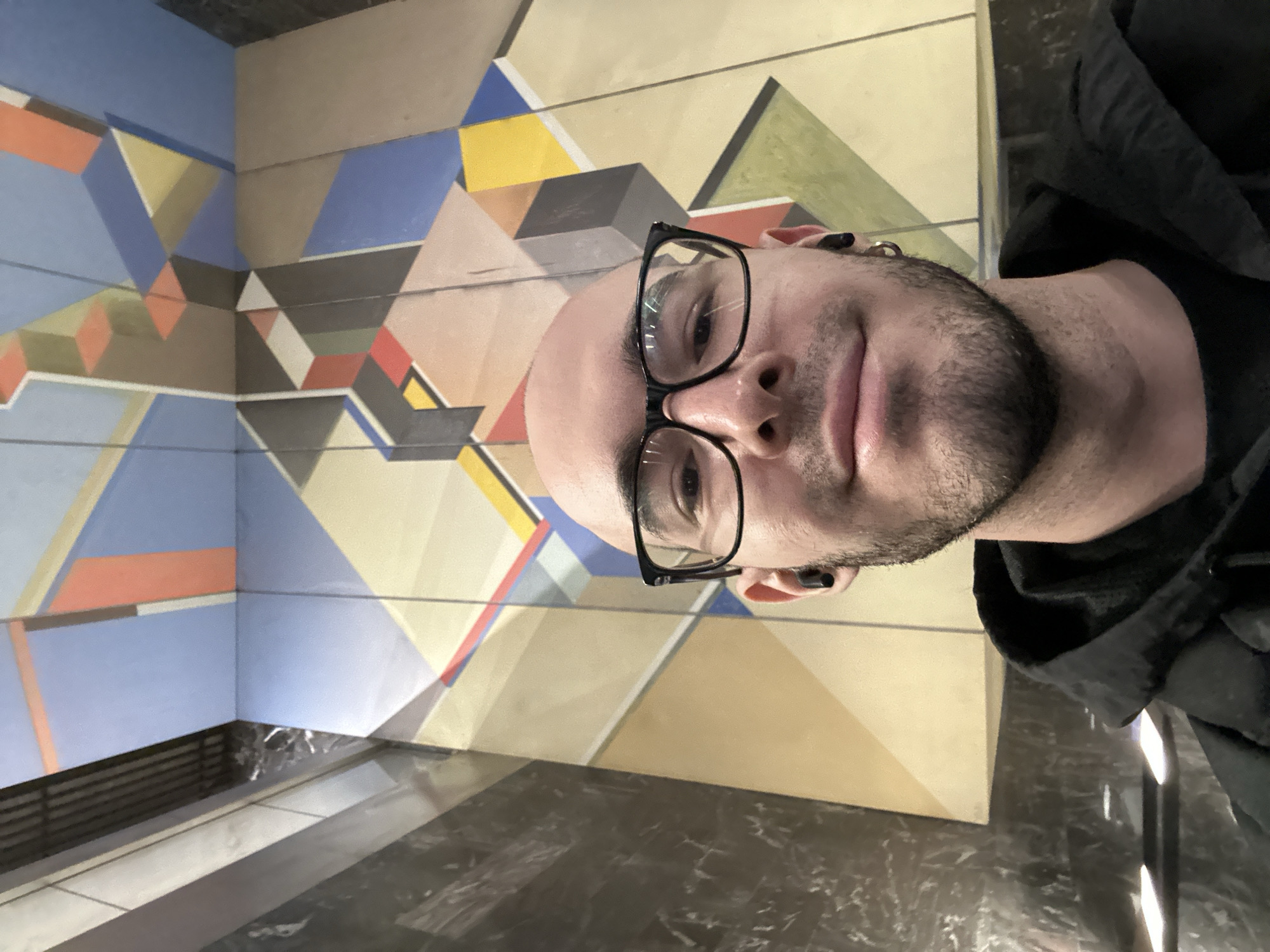 The image shows a man with a shaved head and a beard wearing glasses and a black hoodie. He is facing the camera. The background features a geometrically patterned wall with blue, yellow, and brown colors. The lower portion of the image includes a dark, polished surface that appears to be a floor or a counter. The image was taken in the city of Khimki, Russia.