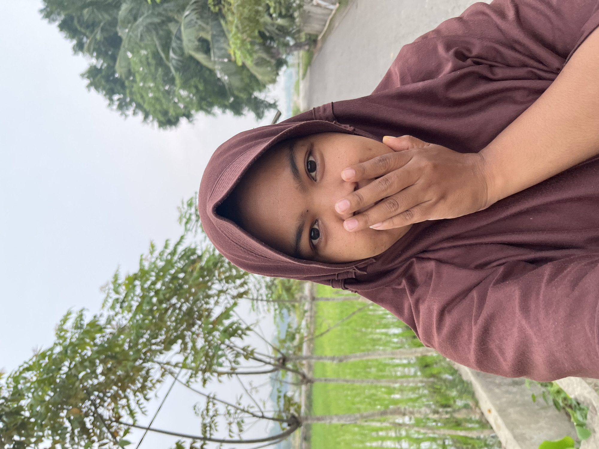 A young woman wearing a maroon hijab stands in front of a rice field, with a road and trees in the background. She is looking at the camera with her hand covering her mouth. The scene is likely in Kruengmate, Indonesia, given the presence of rice fields and tropical vegetation. The sky is overcast, suggesting a cloudy day.