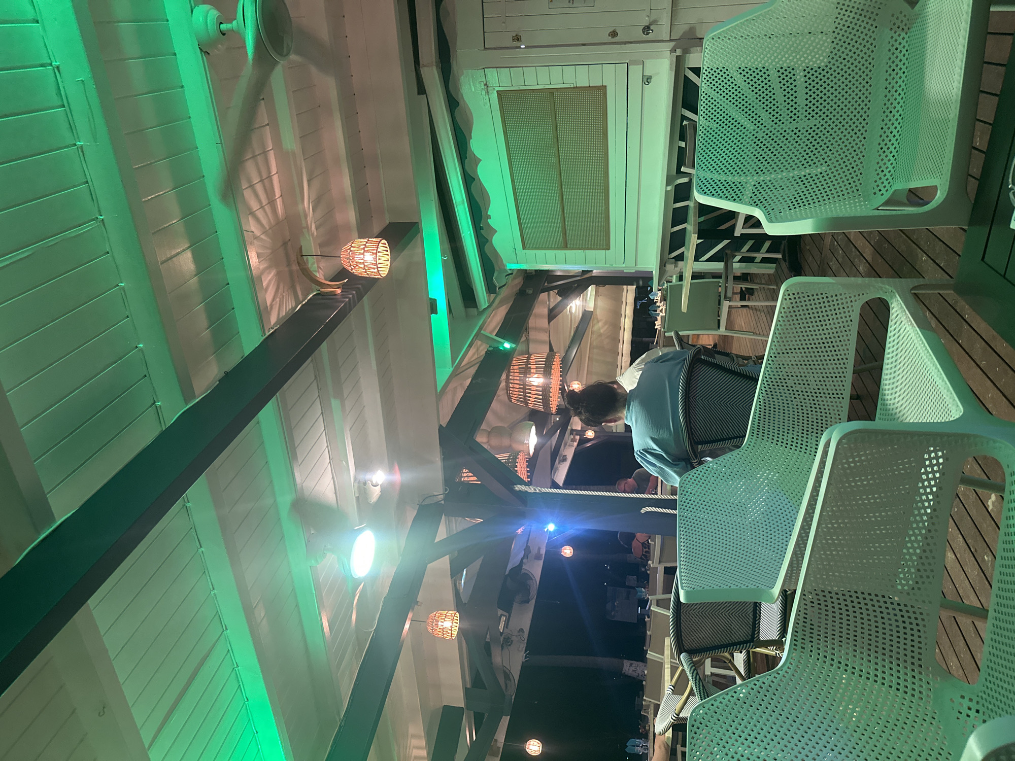The image depicts an outdoor seating area, possibly a restaurant or bar, illuminated with green lighting. Several unoccupied white plastic chairs are arranged on a wooden deck, suggesting a relaxed, casual atmosphere. Other tables are visible further in the background. A person in a blue shirt is seated in one of the chairs. Overhead, the ceiling features a combination of white paneling and dark wooden beams, with wicker lamps and ceiling fans. The overall impression is of a vibrant nightlife setting. It is unknown if the image was taken in Le Gosier, France.