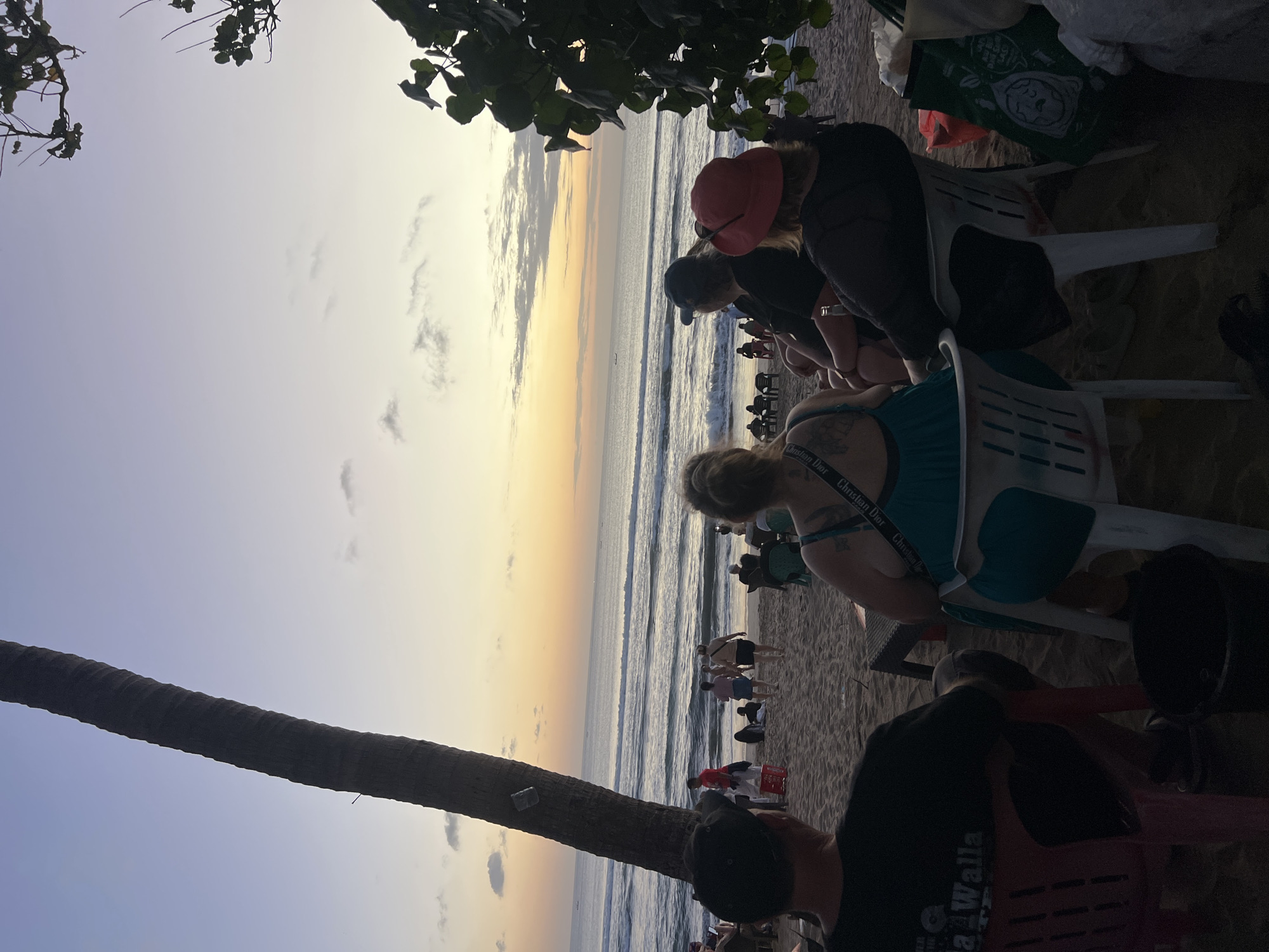The image depicts a beach scene at sunset in Kuta, Indonesia. Several people are seated on chairs on the sand, facing the ocean. The sky is filled with soft hues of orange, yellow, and purple, indicating the sun is setting. Gentle waves roll onto the shore. A tall palm tree stands prominently on the left side of the frame, and foliage from another tree is visible on the right. Other individuals are walking or standing further down the beach, and small boats can be seen in the distance on the water. The overall atmosphere suggests a relaxed evening gathering.