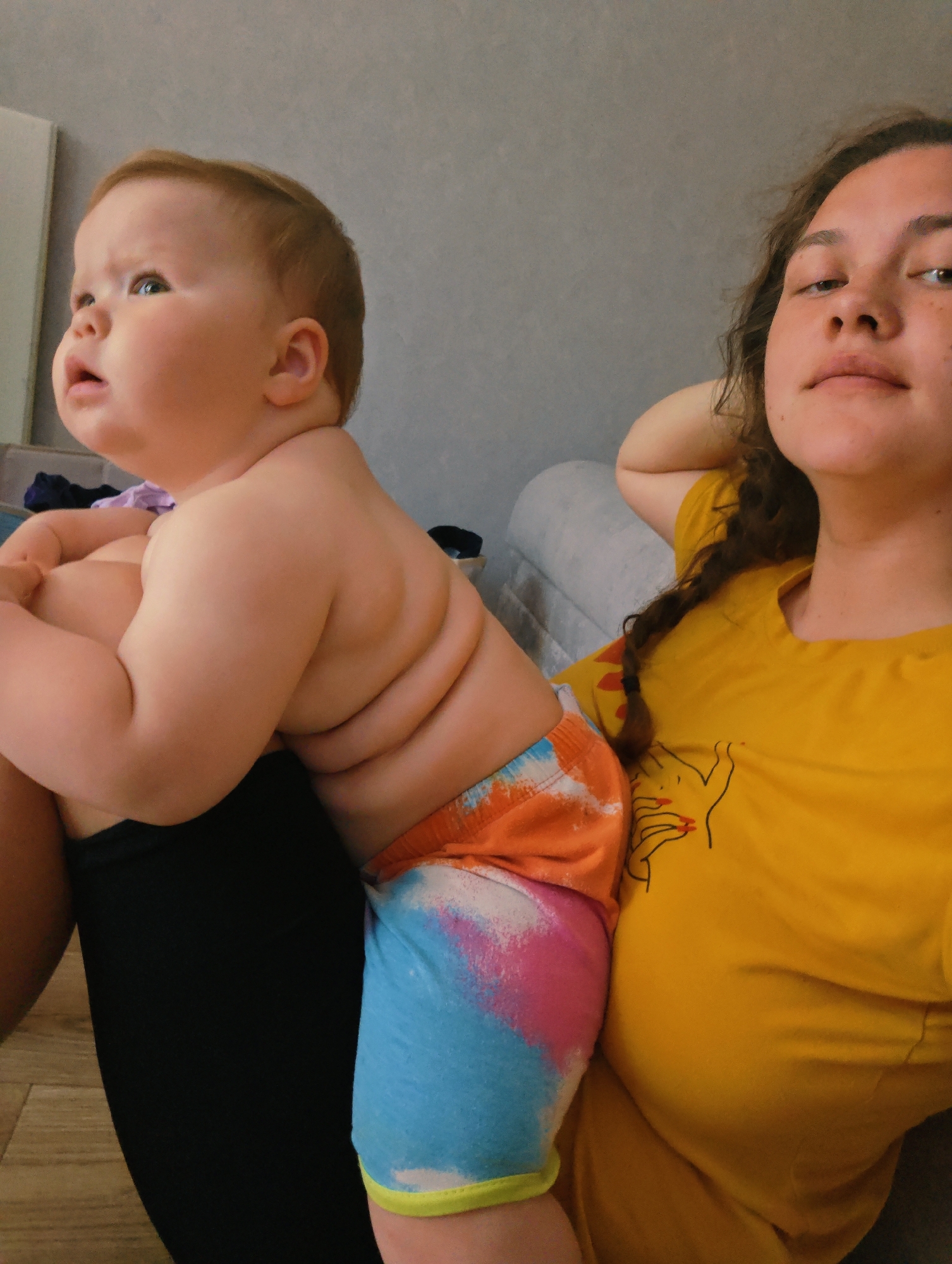 In this media file, a young child is held by a woman. The child, who appears to be a toddler, is sitting on the woman's lap, facing away from the camera and looking upwards. The child has reddish-blonde hair and is wearing brightly colored shorts with a tie-dye pattern of orange, pink, and blue. The woman is wearing a yellow t-shirt with a black line drawing of hands on it. Her hair is dark and curly, and she is resting her head on her hand, looking towards the camera. The background appears to be an indoor setting, possibly a living room, with a grey textured wall and a sofa visible behind them. The lighting suggests it could be daytime. The overall mood seems relaxed and intimate, capturing a moment between a mother and child. There is no visible text in the media file.
