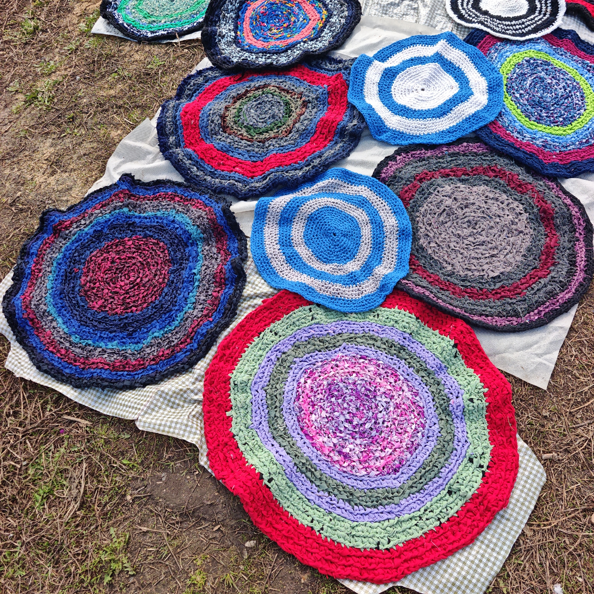 A ground-level shot shows a variety of circular crocheted rag rugs displayed on a white cloth and a checkered fabric outdoors, possibly for sale in an unidentified location. The rugs exhibit a range of colors, including red, blue, purple, green, and gray, often arranged in concentric circles. The setting is natural, with grass and scattered branches visible around the edges of the cloths, indicating an outdoor environment. The scene appears devoid of human subjects.