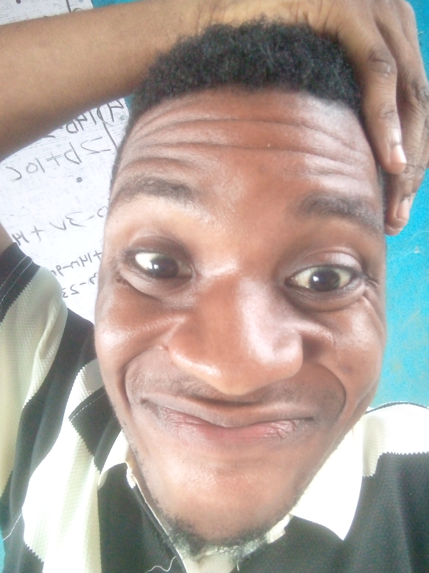 A close-up selfie depicts a man with dark skin and short, curly black hair. His eyes are brown, and his expression includes a slight smile. He is wearing a black and white striped shirt. His right hand is raised and touching his forehead. Behind him, there appears to be a white board with writing on it and a blue wall or surface. The location is Unknown, Unknown.