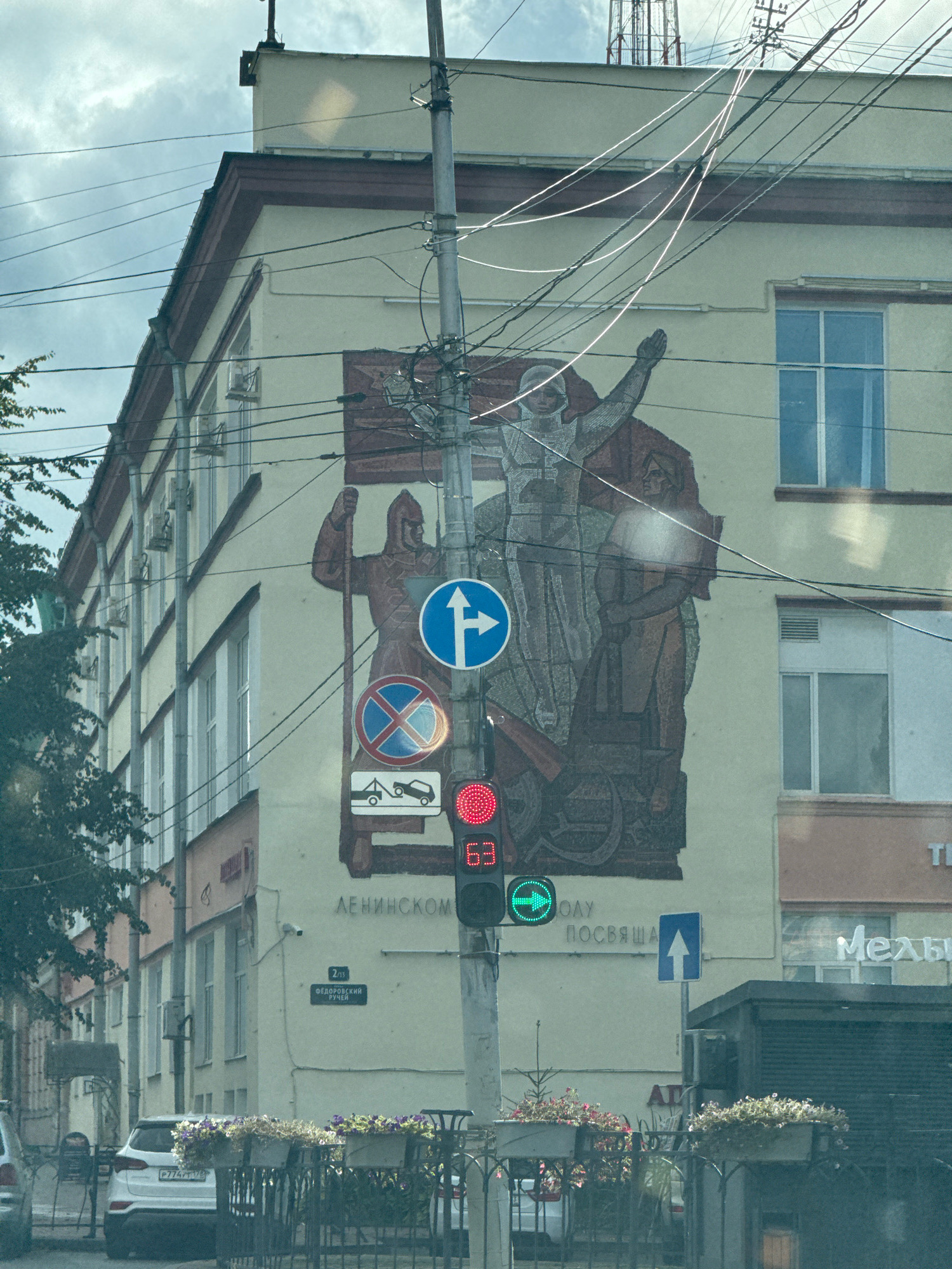 The image captures a street scene in Veliky Novgorod, Russia, focusing on the facade of a building adorned with a large mural. The mural depicts stylized figures, possibly representing historical or allegorical themes, with a central figure raising an arm. Power lines crisscross the building and the mural, obscuring parts of the artwork.
In front of the mural, a traffic light is visible, displaying a red light at the top and a green circular arrow on the bottom light, indicating an allowed turn. Road signs are also present: a blue circular sign with a white arrow pointing right, a red circular sign with a blue border and a red diagonal cross, and a white rectangular sign showing a tow truck towing a car.
Below the mural, text in Cyrillic is partially visible, reading "ЛЕНИНСКОМУ" and "ПОСВЯЩА." A street sign indicates "2/19 ФЕДОРОВСКИЙ РУЧЕЙ."
On the left side of the image, a white car is parked. The foreground shows a black metal fence with potted plants overflowing with flowers. The overall environment suggests an urban setting under a cloudy sky.