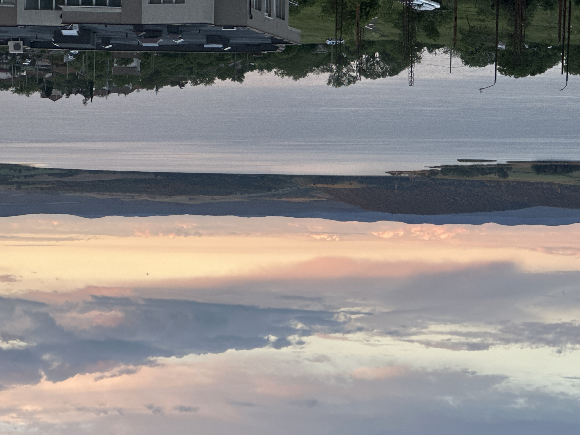 The image depicts a landscape divided horizontally into distinct layers. The bottom portion of the image shows a cloudy sky with hues of gray, blue, and pink, suggesting either sunrise or sunset. Above the sky, there is a landscape composed of dark, rolling mountains, followed by a strip of green land. Above this land, there is a body of water that appears calm. The top portion of the image is a reflection of buildings, trees, and electrical poles on the water's surface. The buildings appear residential, with cars parked nearby. The overall scene suggests a tranquil environment with elements of both nature and human settlement, possibly near a lake or reservoir.