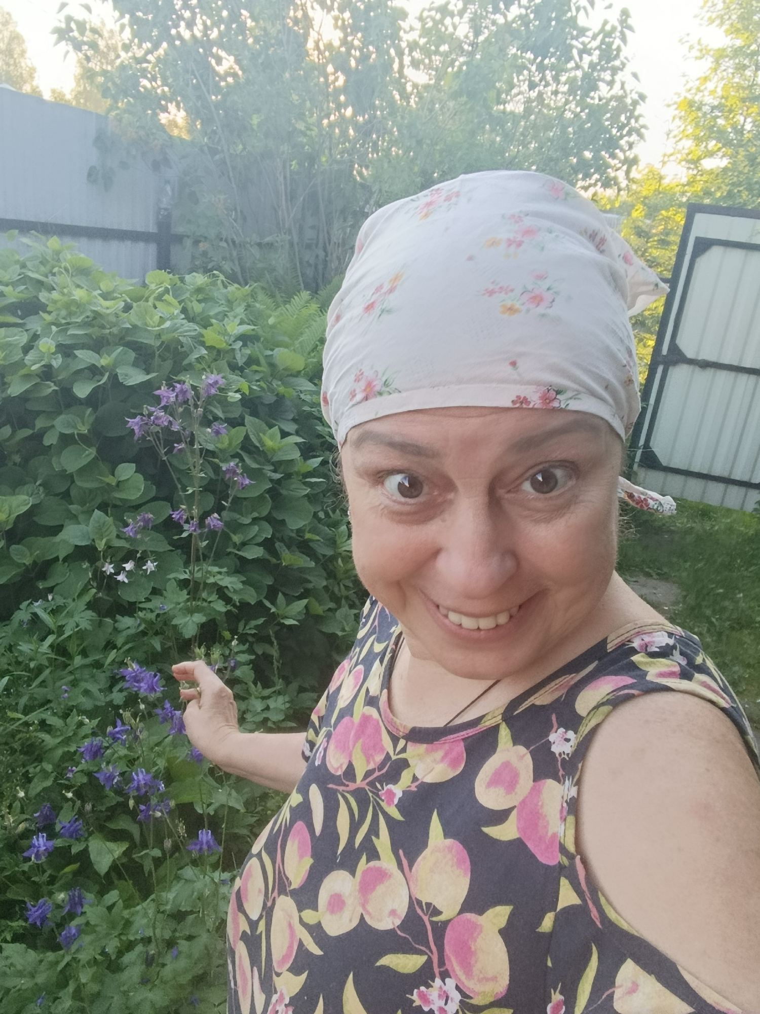 An adult female, likely middle-aged, is depicted in a close-up outdoor photograph taken in the city of Верея, Russia. She is smiling broadly and looking directly at the camera. She wears a light-colored headscarf adorned with pink and yellow floral patterns, and a dark-colored sleeveless top featuring a pattern of pink and yellow peaches or fruits with white blossoms. Her right hand, with a pale complexion, extends towards a cluster of purple, bell-shaped flowers (likely Aquilegia or Columbine) located to her left. Behind her and the flowers is dense green foliage, consistent with a garden environment, featuring various leafy plants. In the background, a light-colored corrugated metal fence is visible on both the left and right sides. Above the fence and foliage, treetops with green leaves are silhouetted against a bright, illuminated sky, indicating an outdoor, daytime setting. Sparse grass is visible on the ground behind her to the lower right.