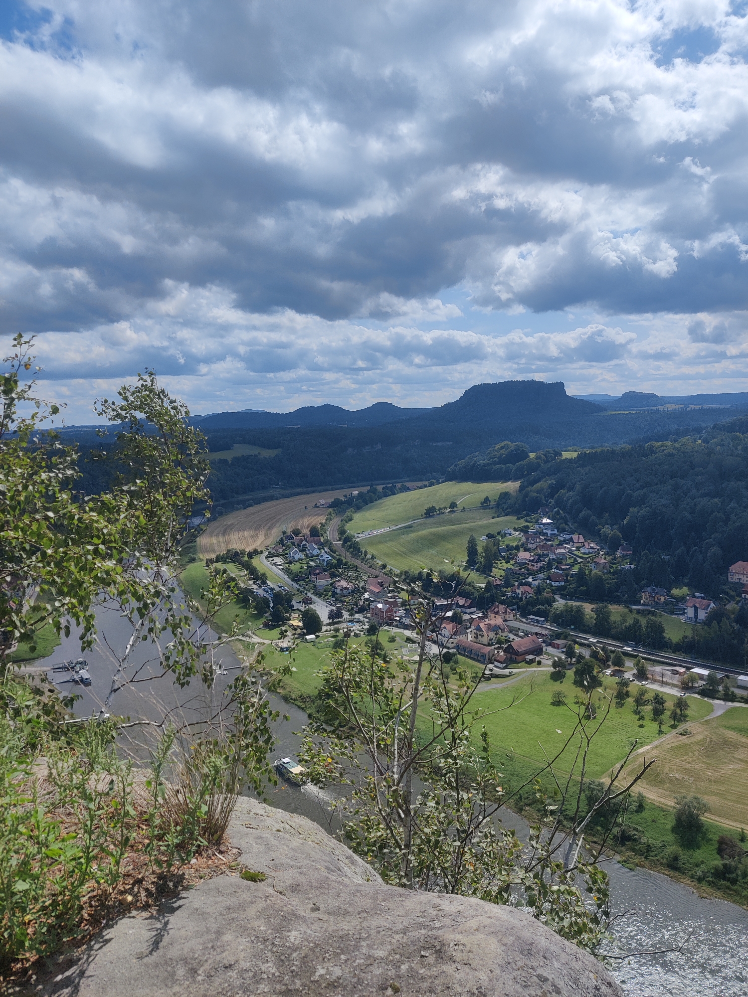 An aerial, high-angle, full shot captures a panoramic vista of the Elbe Sandstone Mountains in Lohmen, Germany, under a cloudy sky. In the foreground, a large, grey boulder is partially covered with green vegetation and dry grasses. A body of water, appearing to be the Elbe River, occupies the lower left of the frame, with several boats docked along its bank.
The middle ground features a verdant landscape with a scattering of buildings forming a small town, nestled between the river and rolling hills. The town consists of houses with red-tiled roofs and a church. A train track runs parallel to the river, with a train visible. Fields of varying shades of green and yellow agricultural land are interspersed throughout the scene.
The background is dominated by the majestic, tree-covered mountains characteristic of the Elbe Sandstone region. Their silhouettes stand against the dramatic, overcast sky, which is a mix of dark grey and white clouds, with patches of blue sky peeking through. The overall impression is one of natural beauty and a peaceful rural setting.