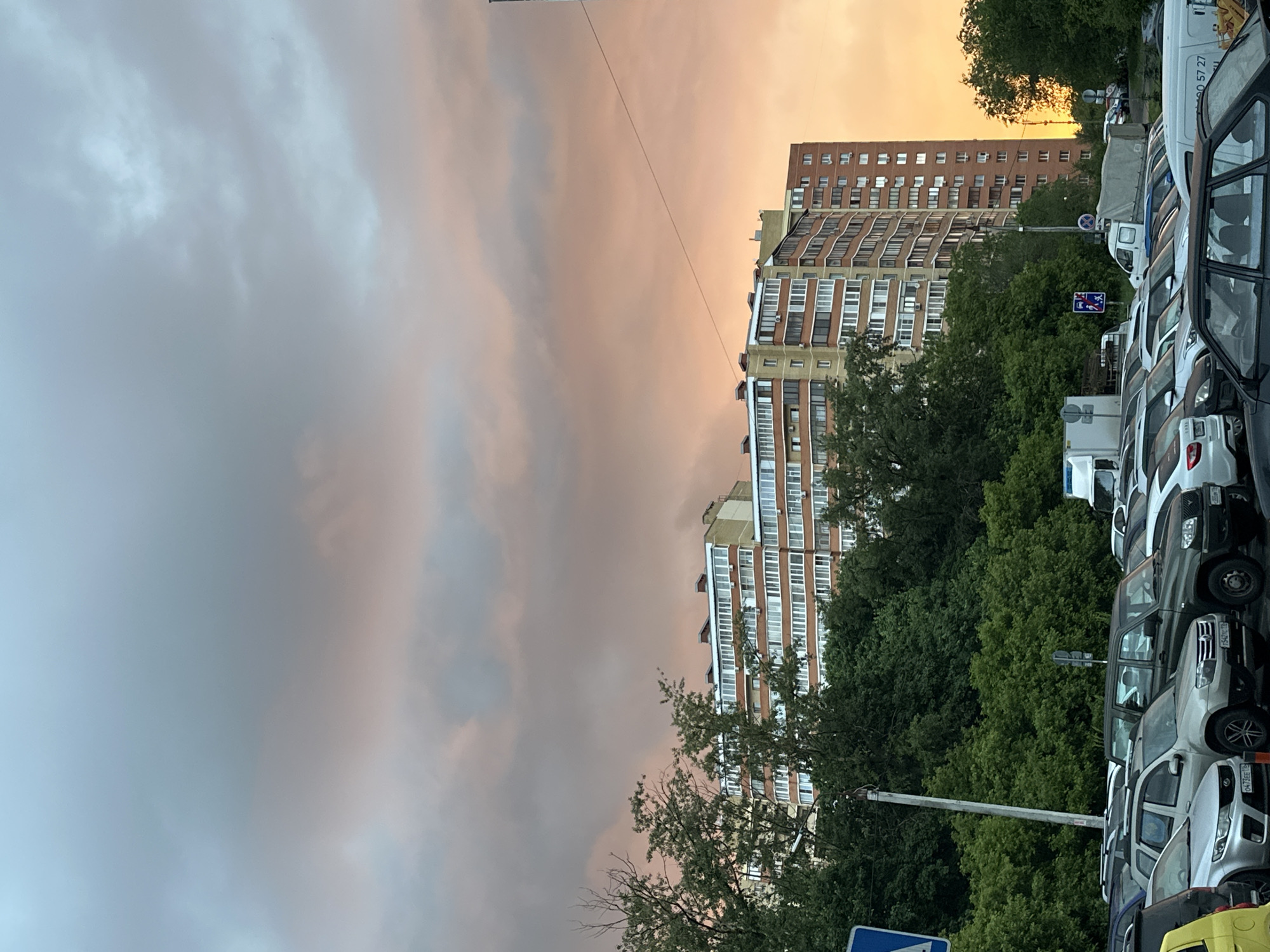 The image, captured in Kazan, Russia, presents an urban landscape at either sunrise or sunset, characterized by a striking sky. The upper and left portions of the frame are dominated by a cloudy sky, transitioning from soft grey and pale blue on the left to vibrant hues of orange, pink, and yellow towards the right, indicating the low angle of the sun. A thin power line diagonally traverses this colorful expanse.
Below the sky, on the right side, stand multiple multi-story residential buildings, their facades marked by numerous windows and balconies. Some structures, made of brick or similar materials, are partially hidden by a dense expanse of green trees. One building exhibits a distinctive wavy architectural design.
The foreground on the right features a long line of parked vehicles, including various cars, vans, and light trucks, many of which are white. Details like license plates are visible; for instance, "116 RUS" is clearly seen on multiple vehicles, confirming the location as the Republic of Tatarstan. One plate specifically reads "007EE 116 RUS". A white van also displays a partial phone number, "0 57 27," along with other illegible Cyrillic text. Traffic signs, including a blue square sign with a white arrow and a blue circular sign, are present amidst the foliage and vehicles. A utility pole also stands within the scene. No specific activities or people are discernible, presenting a tranquil urban moment.