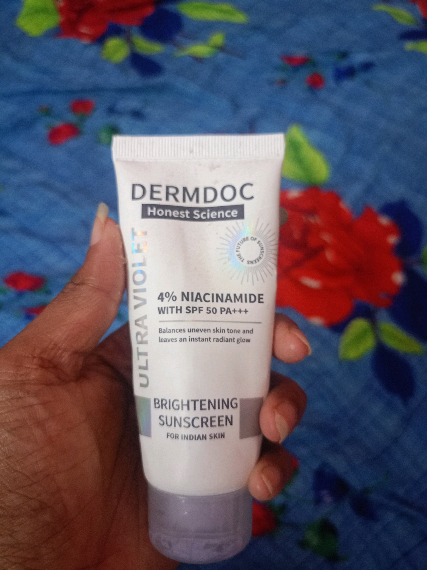 In Chenkavila, India, a person's hand holds a white tube of DERMDOC Brightening Sunscreen, with the text "ULTRA VIOLET 4% NIACINAMIDE WITH SPF 50 PA+++" visible on the label. The background is a blue patterned fabric with red and green floral motifs.