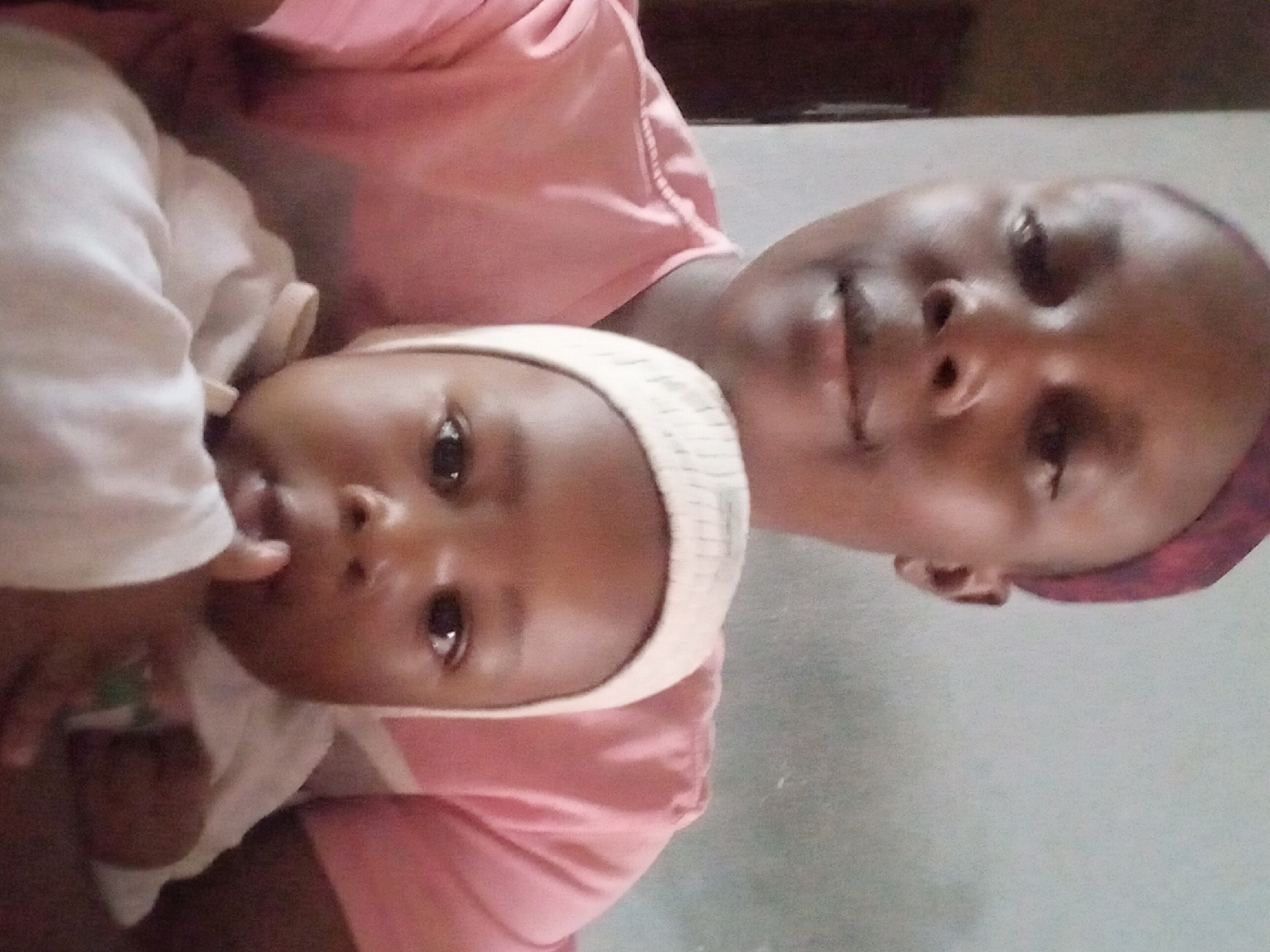 A medium shot depicts a woman holding a baby. The woman is wearing a pink shirt and has short hair covered with a purple headscarf. She is looking at the camera with a slight smile. The baby is wearing a white hat and shirt, looking directly at the camera with a hand near its mouth. The background is blurry, and the location is identified as Sabon Tasha, Nigeria.
