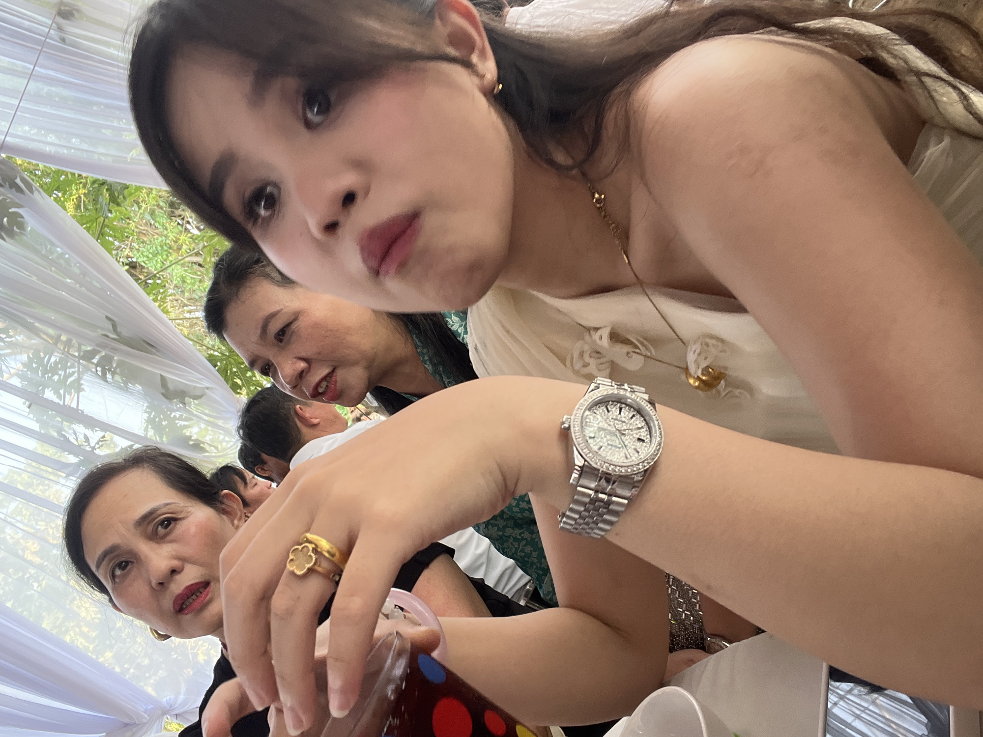 This close-up, low-angle photograph captures a social gathering in Bình An, Vietnam. The primary subject is a young woman in the foreground, looking directly at the camera with a slightly playful or pouty expression. She has long, dark hair and is wearing a light-colored garment, possibly off-shoulder, accessorized with a delicate gold-toned necklace. On her left wrist, she wears a silver-toned wristwatch featuring a diamond-studded bezel and a sparkling dial, while a gold-toned ring with a clover-like design adorns her right ring finger. She holds a transparent cup decorated with colorful circular patterns (red, blue, green).
Behind the main subject, two older women are partially visible. One woman, positioned in the mid-ground, appears to be smiling subtly. Another older woman, lower and to the left, has a more neutral expression and wears red lipstick. The head of a man is also partially visible between the figures.
The setting is an open-air or semi-outdoor event, suggested by the presence of white fabric draped overhead, resembling a tent or canopy. Lush green foliage is visible in the background, indicating a daytime event. White ceramic cups or bowls are also visible on a surface in the lower right, suggesting a meal or refreshments are being served. The image conveys a sense of intimate observation within a festive or communal atmosphere. There is no visible text.