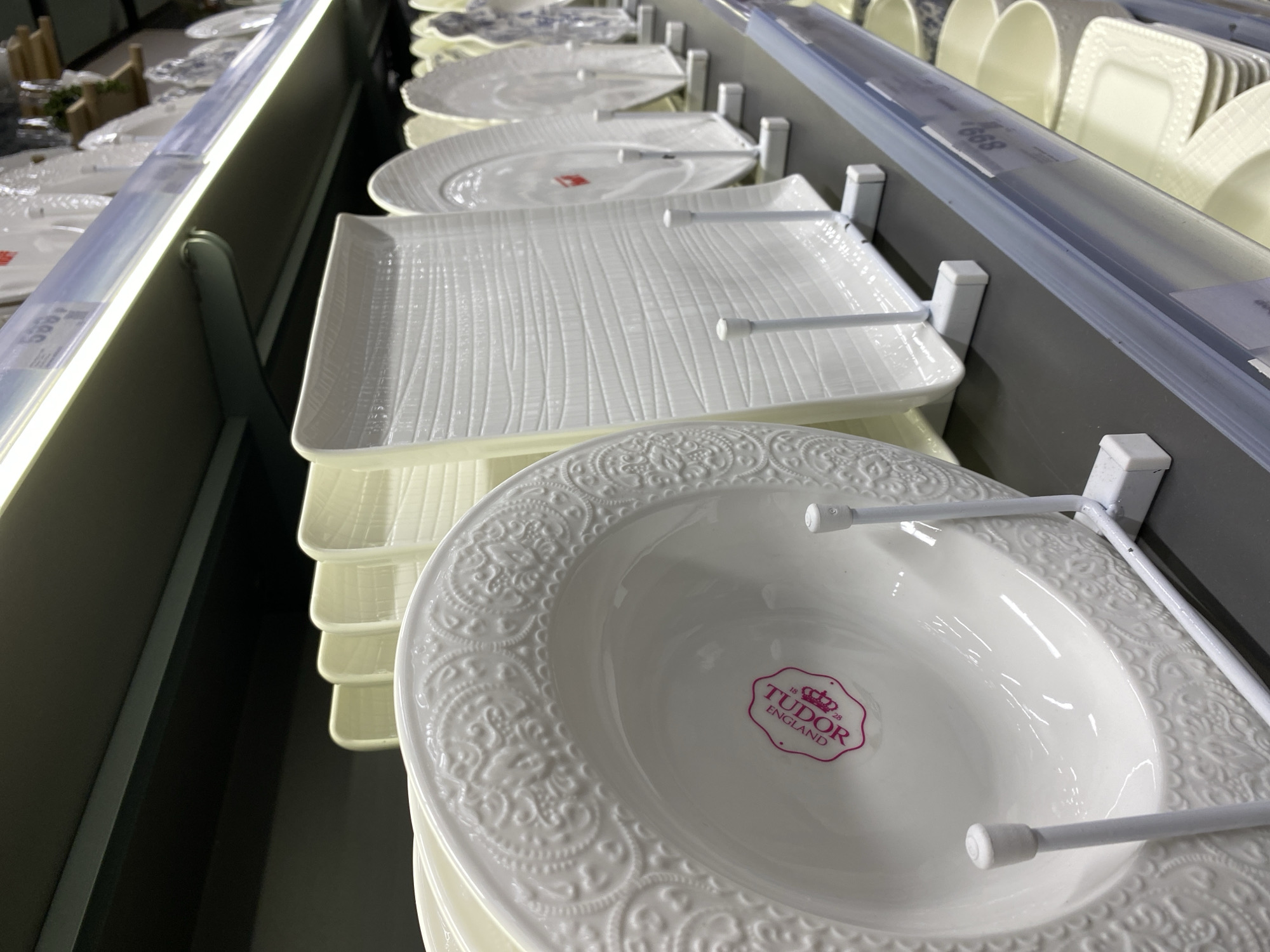 The image shows a shelf of white plates and platters for sale, likely in a store in Murino, Russia. The brand "Tudor England" is visible on a stack of decorative serving bowls at the front of the shelf. Other plates include rectangular serving dishes with a ridged texture. The plates are displayed on metal racks. The lighting is bright, possibly from shelf lighting. The background shows a partial view of other dishware and store fixtures.