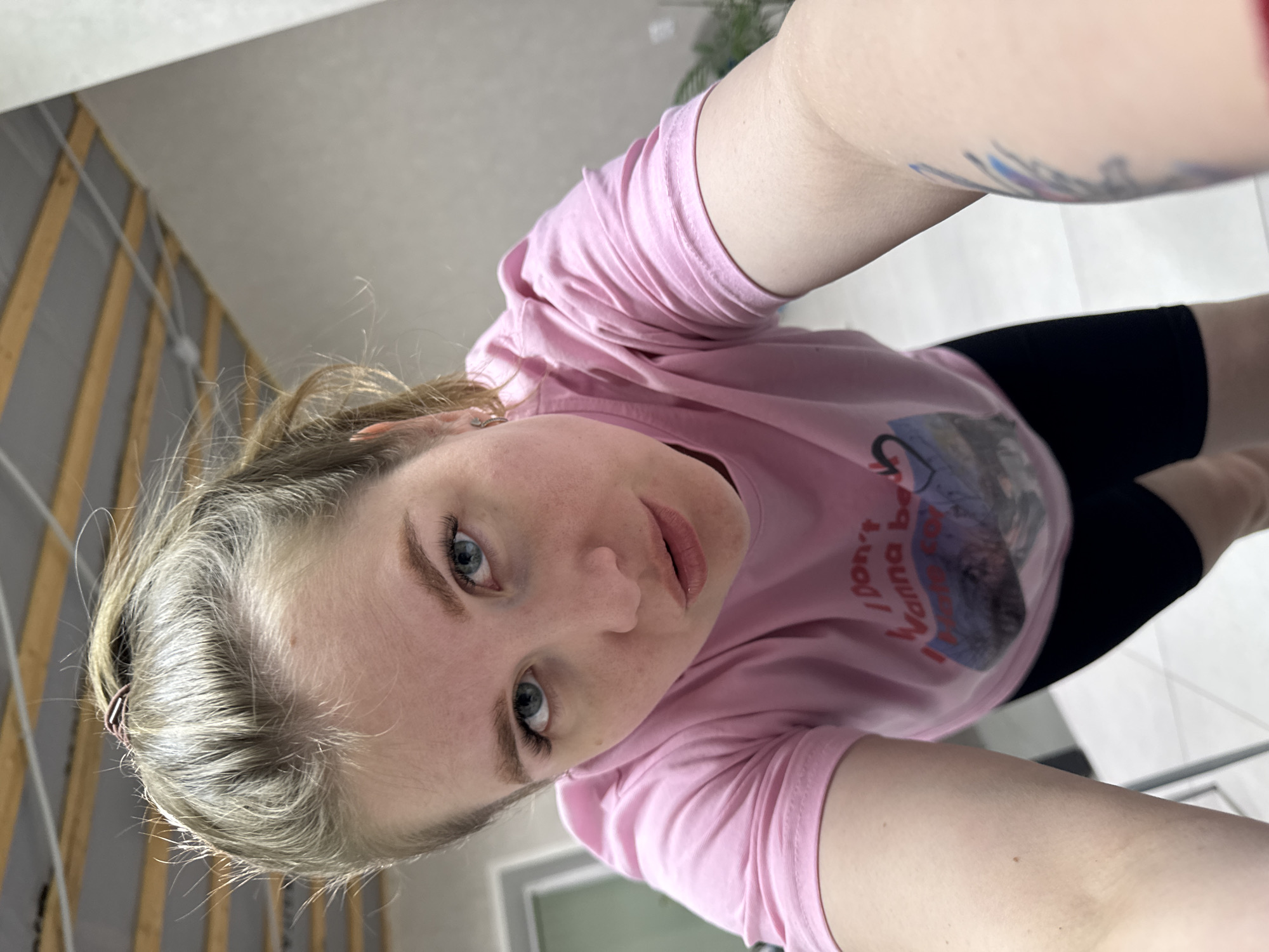 A woman is taking a selfie. She has light skin, blue eyes, and blonde hair pulled back. She is wearing a pink t-shirt and black shorts. A tattoo is visible on one arm. The background includes a wall with visible wiring and a tiled floor. The location is Gornyi (Dvurechenskoe S/P), Russia.