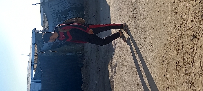 The image displays a male individual walking right-to-left on an unpaved, light brown dirt path in Khan Yunis, Palestinian Territory. He is dressed in a black tracksuit featuring red chevron patterns across the chest and red stripes down the outer seams of the pant legs; a white Nike swoosh logo is visible on the upper left chest. He wears brown sandals and a light brown backpack. His head is turned towards his right, looking over his shoulder. A ring is visible on his left ring finger. Long, distinct shadows of his figure are cast on the ground, indicating direct sunlight from a low angle. In the background, several makeshift structures are visible, constructed from dark tarpaulins, blankets, and possibly other salvaged materials. One structure includes a light blue tarp section forming part of its roof. Arabic script and white floral patterns are discernible on a dark blanket used as a wall for one of these structures. The sky above is clear and light blue. The ground surface shows visible footprints.