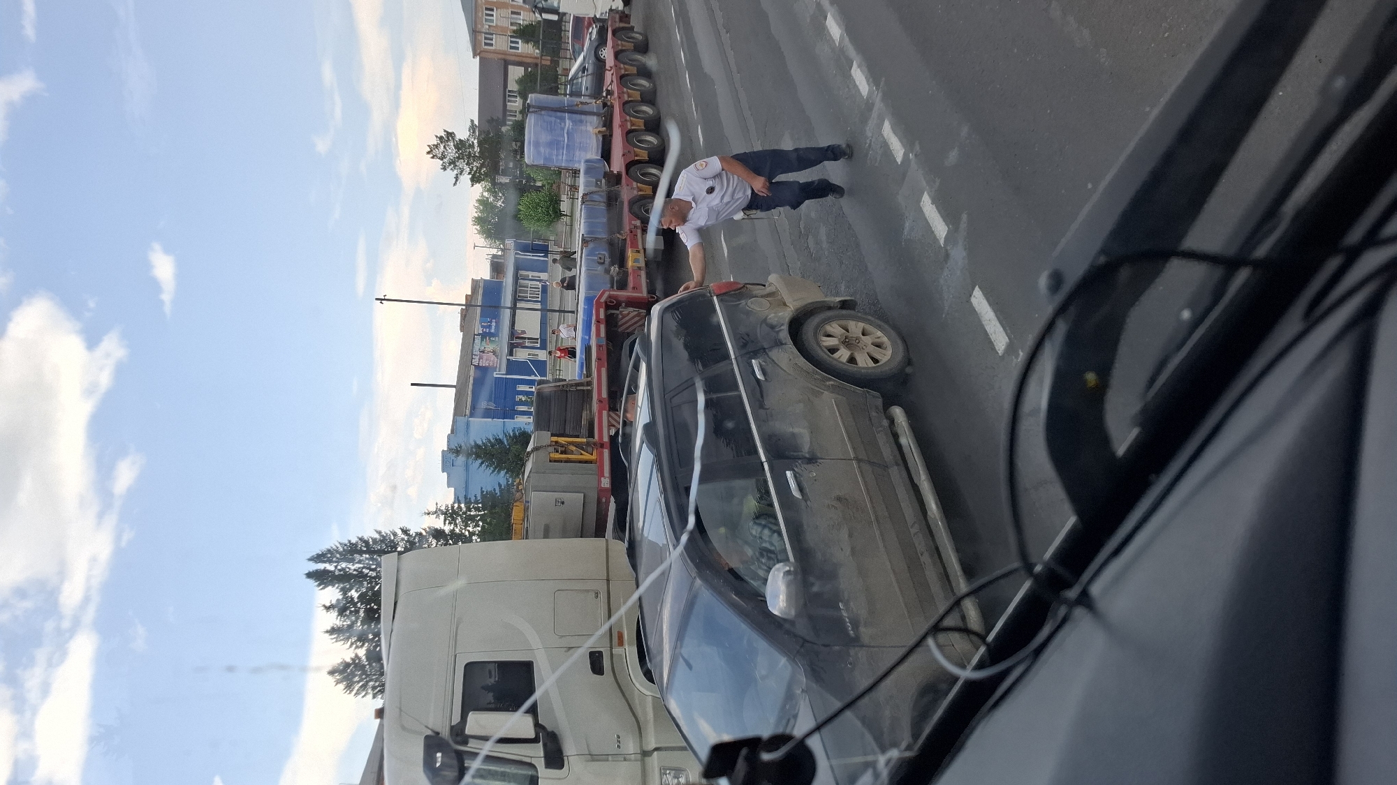 In Nizhny Ingash, Russia, a man in a white collared shirt and dark pants, likely a traffic controller or law enforcement officer, leans his right hand on the rear passenger door of a dark-colored SUV. The SUV is positioned on a road with white dashed lane markings. Behind the SUV, a long, flatbed trailer, laden with large blue cylindrical objects, is being towed by a white semi-truck. The trailer has multiple axles, indicating it's designed for heavy loads. In the background, a row of buildings lines the street, some painted in blue, with trees visible between them. The sky above is partly cloudy, with a mix of blue sky and white clouds. The overall scene appears to be an urban or semi-urban environment, possibly during daylight hours. The SUV appears dusty, suggesting it may have been driven on unpaved roads.