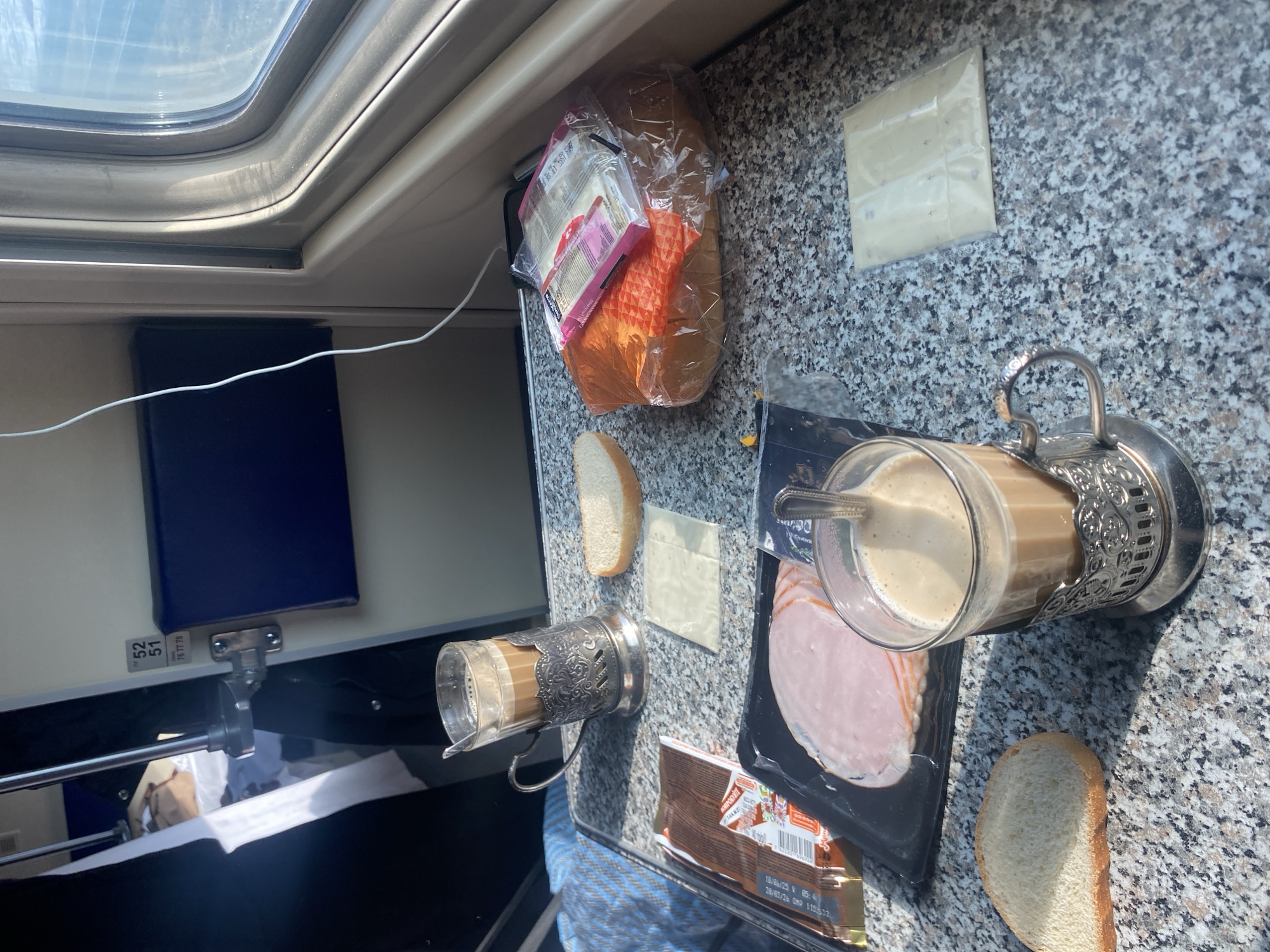 The image depicts the interior of a train compartment, situated in a train known to be in or passing through Kotelnich, Russia. A speckled gray and white tabletop, consistent with a fold-down train table, occupies the foreground. On the table, various food items are arranged: two glasses containing a light brown, foamy beverage, likely coffee with milk, are seated in ornate silver-colored metal podstakanniki (glass holders), each containing a spoon. Three separate slices of white bread are present on the table surface. A plastic-wrapped package of sliced pink luncheon meat or ham is visible, along with two individual, unwrapped slices of light-colored cheese. Additionally, a clear plastic bag contains a loaf of orange-colored bread, topped with a smaller plastic package of cheese slices. A brown rectangular package, possibly chocolate, with Russian text and an expiration date is also on the table. A white charging cable runs across the table, extending towards a window on the right side of the frame, which shows a bright, possibly sunlit exterior. To the left, a blue plaid blanket or fabric covers a surface. A partial view of an interior doorframe is visible in the background, displaying black numbers "52" and "51" vertically, with "76 77 78" below, and a dark blue panel. Behind the doorframe, a glimpse of an interior with a white-linened bed and headrest is apparent. No individuals are visible.