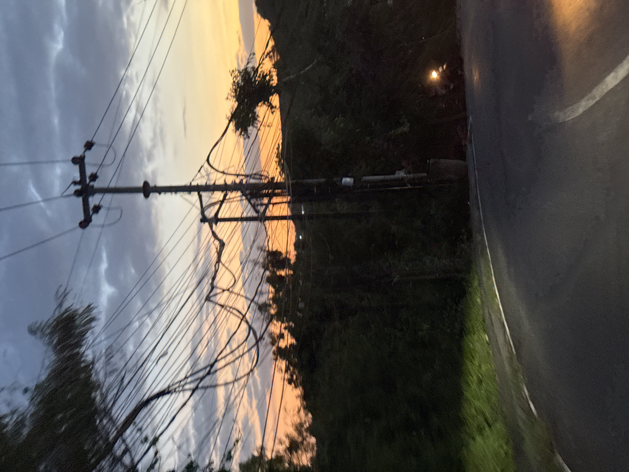 The image shows a roadside scene in Kuta Selatan, Indonesia at dusk. The sky is a mix of grey and orange, indicating a sunset or sunrise. A utility pole with numerous power lines dominates the left side of the frame. Dense, dark foliage lines the side of the road. A paved road curves into the distance on the right. A single light is visible in the background. No people are visible.