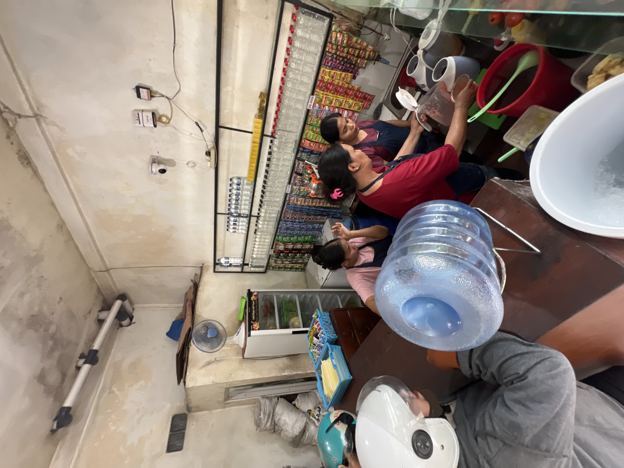 The image shows the interior of a small retail establishment in Jimbaran, Indonesia. Two women wearing aprons stand behind a counter, preparing food or drinks; one wears a red shirt, while the other wears a navy apron. Shelves lining the back wall display a variety of bottled beverages and packaged goods. In the foreground, a blue plastic water jug sits on the counter next to a large white bowl that appears to hold ice. A person wearing a grey jacket and a white and teal motorcycle helmet sits on the far right in the foreground. The shop appears to be located in an older building, with visible wear on the walls. A small fan is mounted on the wall in the lower left.