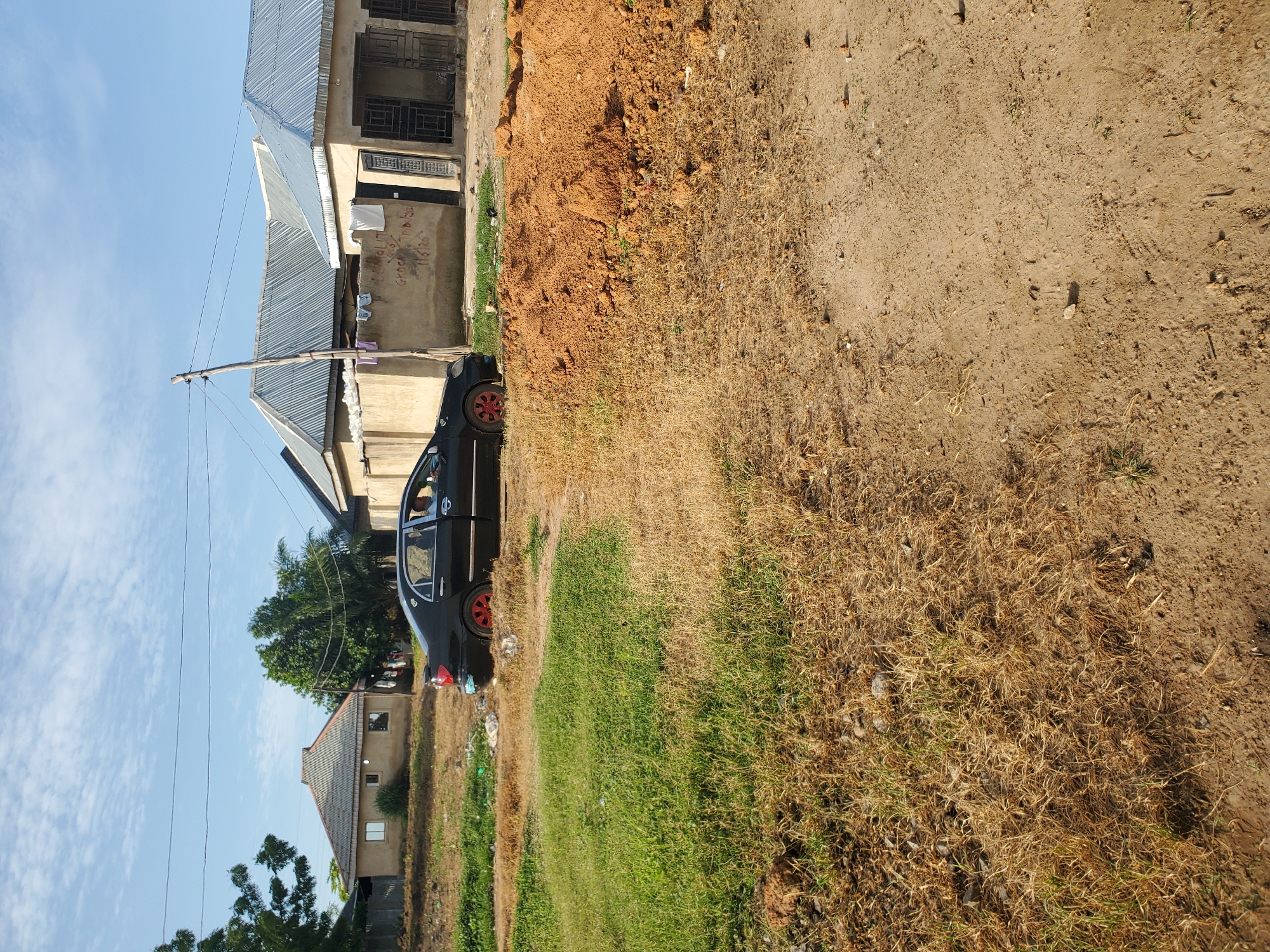This image depicts an outdoor residential scene in the city of Winters, United States of America, under a clear sky with scattered white clouds. The foreground consists of uneven ground, primarily covered with a mix of dry, brownish-yellow grass and patches of green vegetation, leading to a dirt path on the right. A large pile of reddish-brown soil is visible in the mid-right foreground.
A black sedan, resembling a Toyota Corolla, is parked in the mid-ground, facing towards the right. The car features distinctive red-painted wheel rims. The driver's side door is open, revealing a person, likely male, wearing a light blue collared shirt, seated behind the steering wheel.
Behind the vehicle, a low, light-colored concrete wall or fence is visible, with some clothes hanging over it. Graffiti, including text like "DANHOLIT," "GROCA," and "116126," is present on this wall. Beyond the wall and the car, several single-story buildings with corrugated metal roofs are visible. To the far left, a house with a grey ridged roof and light-colored walls features small rectangular windows, partially obscured by green trees. Further right, another building with a similar roof and light-colored walls has dark metal security bars on its windows and doors. A wooden utility pole with overhead wires stands prominently between the car and the buildings on the right. Green trees and dense foliage are scattered throughout the background.