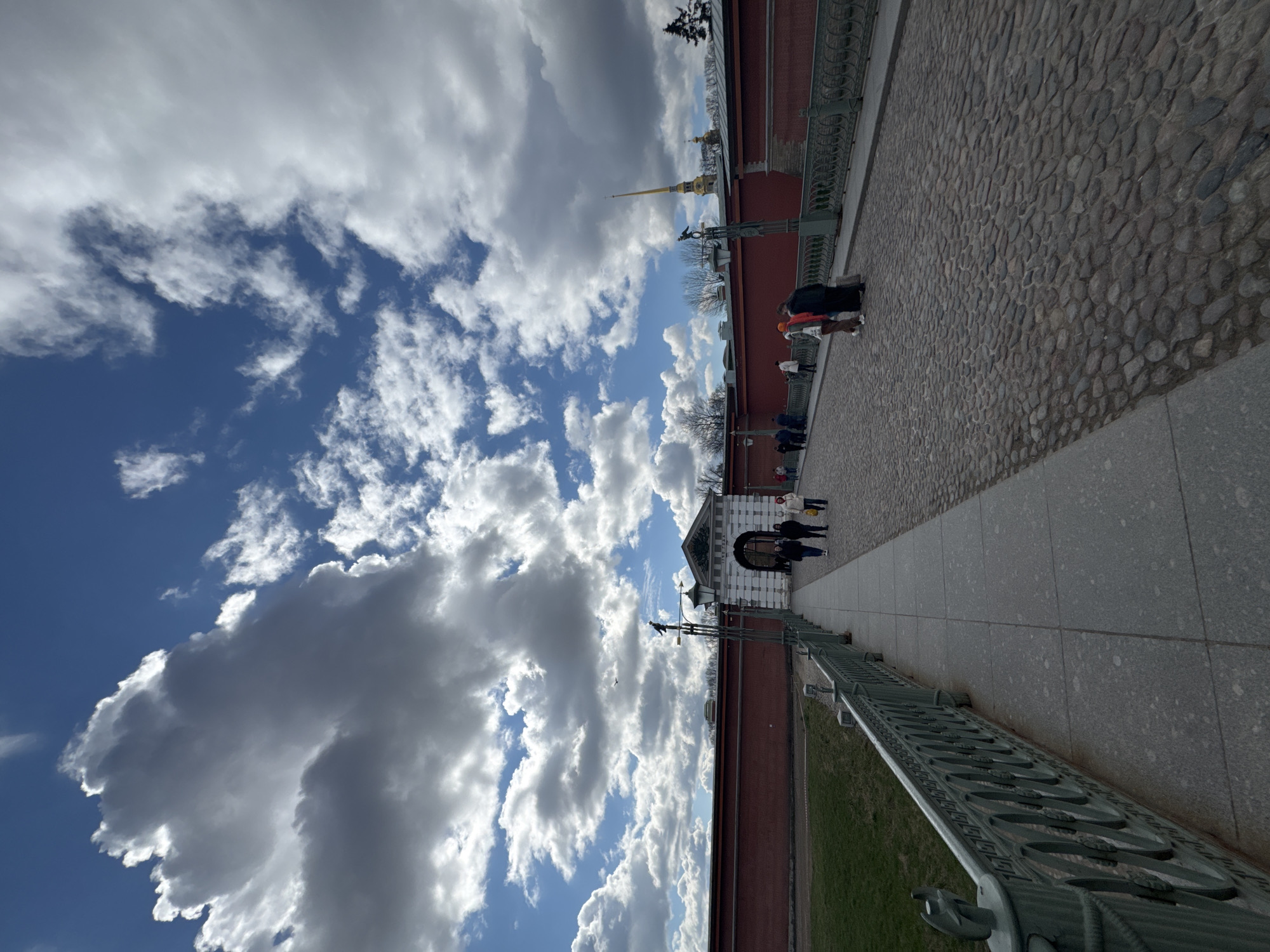 The image shows a view along the wall of the Peter and Paul Fortress in Saint Petersburg, Russia. The sky is blue with puffy white clouds. On the right of the image is a stone pathway and on the right edge, a metal fence separates the pathway from a grassy area. The fortress wall is made of red brick. Several people are walking along the pathway. In the distance, the golden spire of the Peter and Paul Cathedral is visible. The image is taken at an angle.