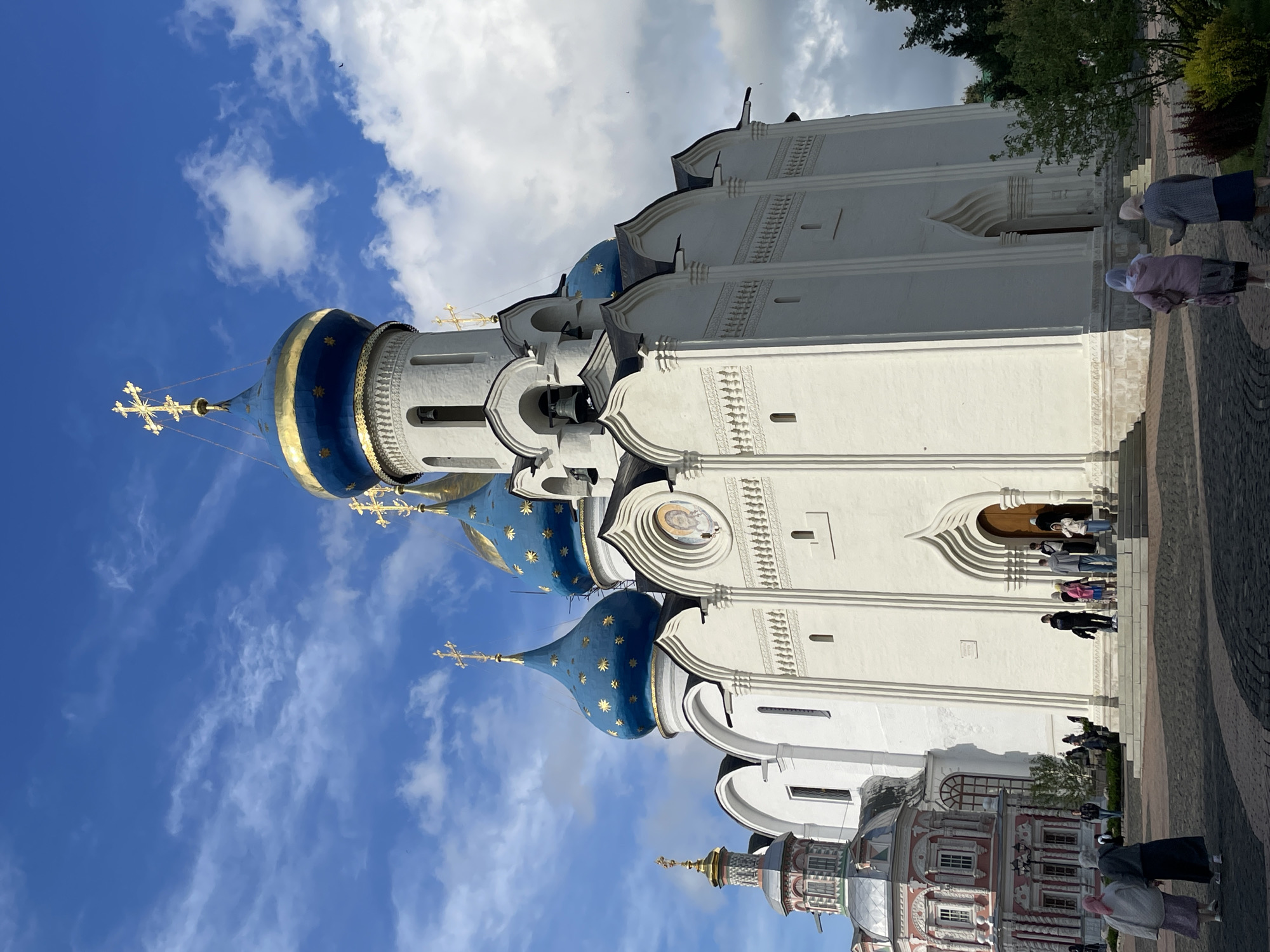 The image shows the Trinity Lavra of St. Sergius in Sergiev Posad, Russia. The main subject is a large white church with multiple blue domes adorned with gold stars and crosses. To the left, a smaller, ornate pink and white building with a silver dome is visible. Several people are walking in the foreground and ascending the stairs to the church entrance. The sky is blue with scattered clouds.