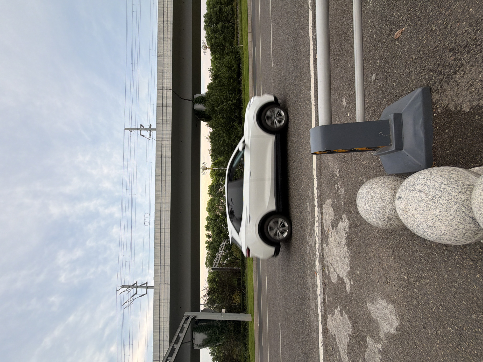 In Jiangjin, China, a white SUV drives on a highway under a viaduct carrying overhead railway lines. The sky is cloudy with streaks of white clouds against a pale blue backdrop. The viaduct's concrete structure is visible, supported by pillars, with the railway tracks and catenary system above. Below the viaduct, a line of lush green trees forms the background. The road is asphalt with white lane markings. In the foreground, a section of a grey metal guardrail with yellow reflectors is visible. To the left of the guardrail, several large, spherical granite bollards are placed on the asphalt. The overall scene depicts a moment of transit on a suburban highway.