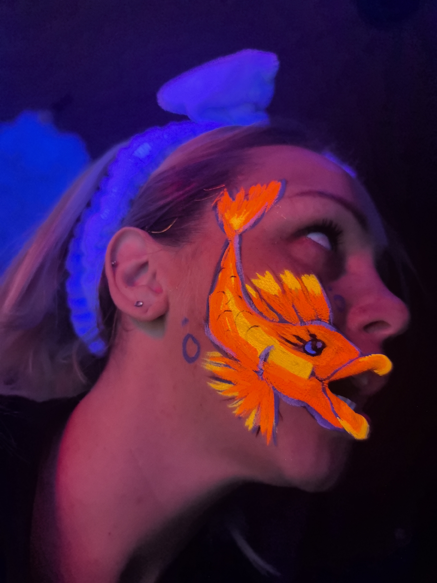 A woman's face is illuminated in a dark environment with neon lighting, creating a vibrant color palette of pinks, oranges, and blues. On her right cheek, there is a stylized face-painted depiction of a fish, rendered in bright orange and yellow hues with a dark outline. The fish appears to be an open-mouthed predatory type. A small, blue circle is visible near her ear. The woman has light hair that is partially obscured by a blue, textured headpiece. Her eyes are slightly upturned, and her mouth is open, possibly in a conversational posture. The background is dark, with nebulous blue shapes. It is unlikely this image is from Russia, given the style and color choice.