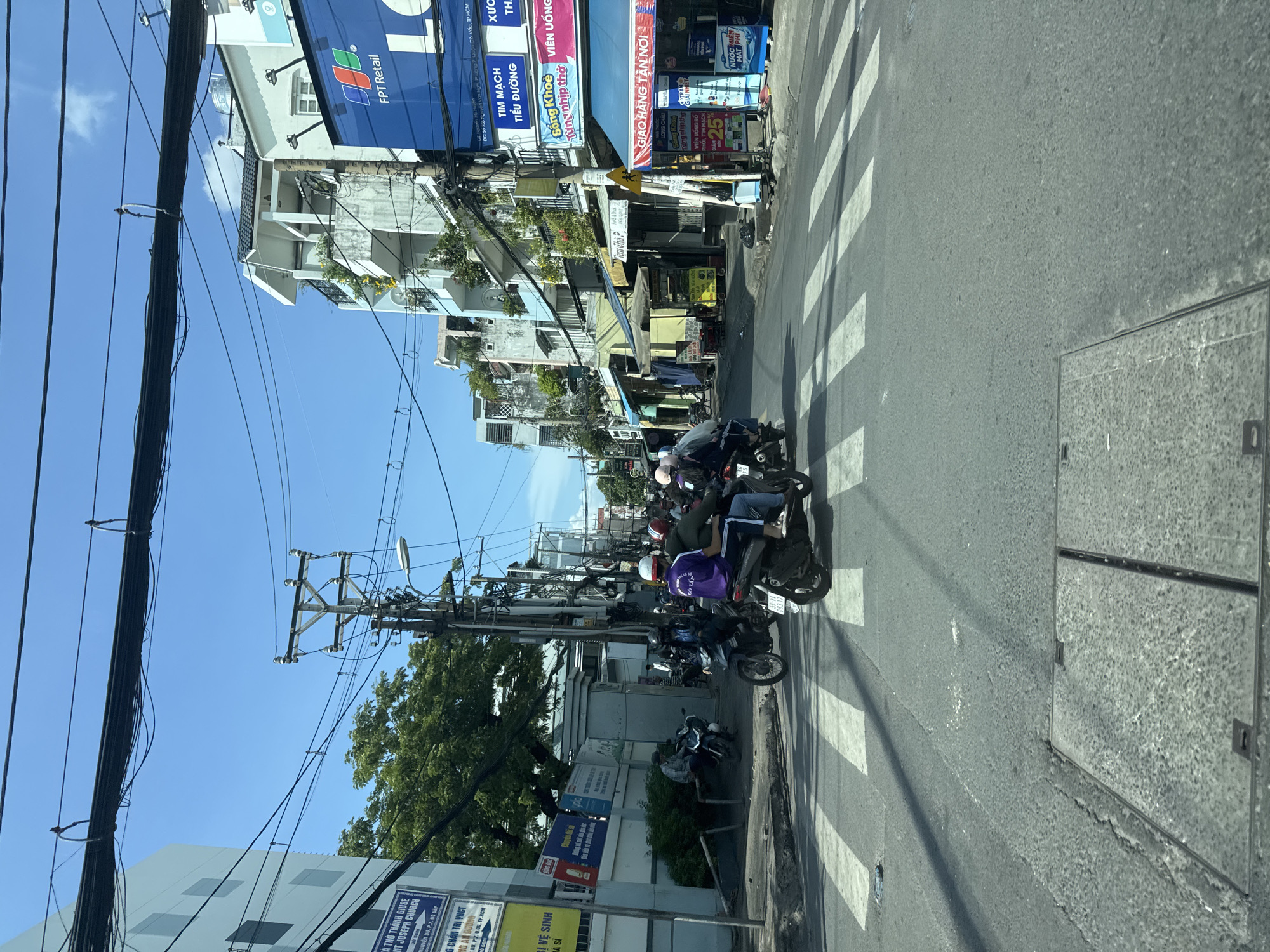 This is a medium shot taken from inside a vehicle, looking out at a street intersection in Thủ Đức, Vietnam. The time of day appears to be daytime with clear, sunny skies. The street is paved and marked with a white pedestrian crossing. Several motorcycles are present, with people riding them, crossing the intersection or stopped on the side. On the right side of the street, there are buildings with businesses and signage, including an FPT Retail store and shops with Vietnamese text. The buildings are of varying heights and architectural styles. Overhead, numerous electrical wires are visible, strung between utility poles. In the foreground, a manhole cover is visible on the road. The overall scene depicts a busy urban street environment.