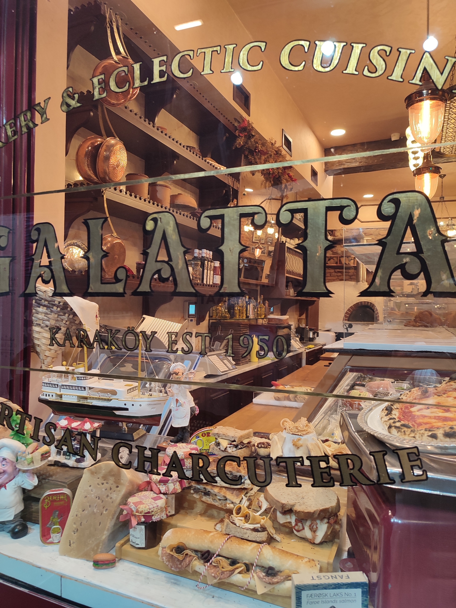 The image shows the storefront of a business named "GALATA" with text above it reading "BAKERY & ECLECTIC CUISINE". The window displays a variety of food items and decorative elements. Prominently displayed are several sandwiches and a pizza. There are also jars of preserves and what appears to be a large wedge of cheese. To the left, a decorative chef figurine is holding a tray with food. Behind the display, shelves are visible with various items, including copper pots, jars, and what looks like a model ship. The interior of the establishment features exposed brick walls, wooden shelves, and ambient lighting from chandeliers. The setting appears to be a delicatessen or a restaurant in Beyoğlu, Turkey, given the name "GALATA" which is a prominent neighborhood in Istanbul, Turkey.