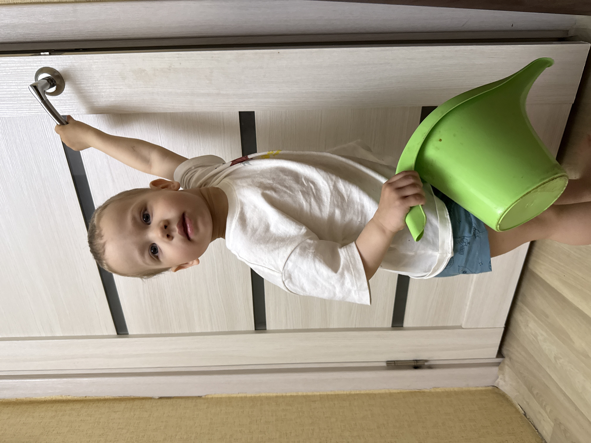 A young child stands near a white door with dark vertical accents, reaching for the door handle with their left hand. They are holding a green watering can with their right hand. The child is wearing a white T-shirt and blue shorts. The room has light-colored walls and flooring. The location is Rusanovo, Russia.