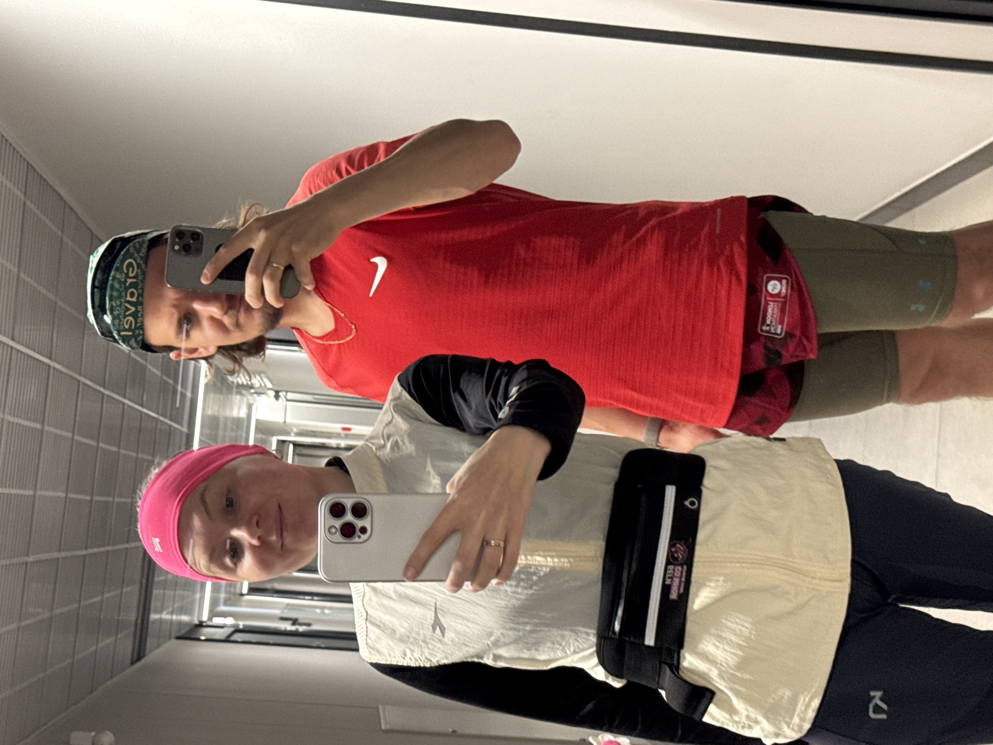 Two people, a man and a woman, are taking a mirror selfie. The man is wearing a red shirt with a white Nike logo, a patterned bandana, olive shorts, and a gold necklace. The woman is wearing a pink headband, a black long-sleeved shirt, a light beige vest, and dark pants. She has a black runner's belt around her waist. The background appears to be a brightly lit public restroom with tiled walls. It is unlikely that the location is Verkhnii Liuk, Russia, as the environment doesn't strongly indicate it.